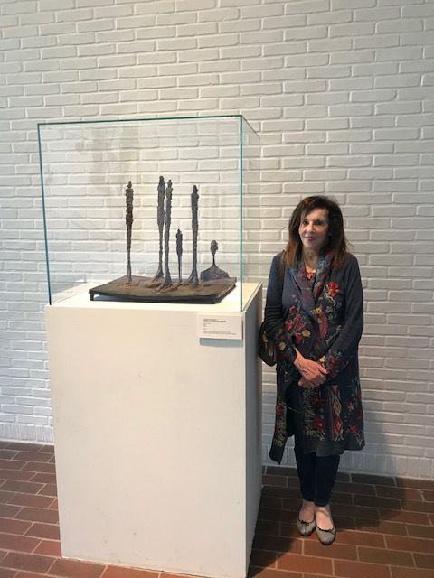 Woman standing next to a glass display case with sculptures inside, against a brick and white wall background.