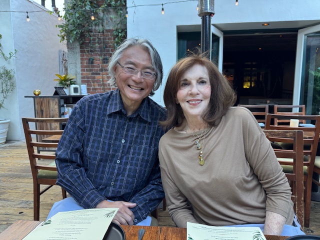 Two smiling people sitting at a table outdoors, with chairs and a building in the background.
