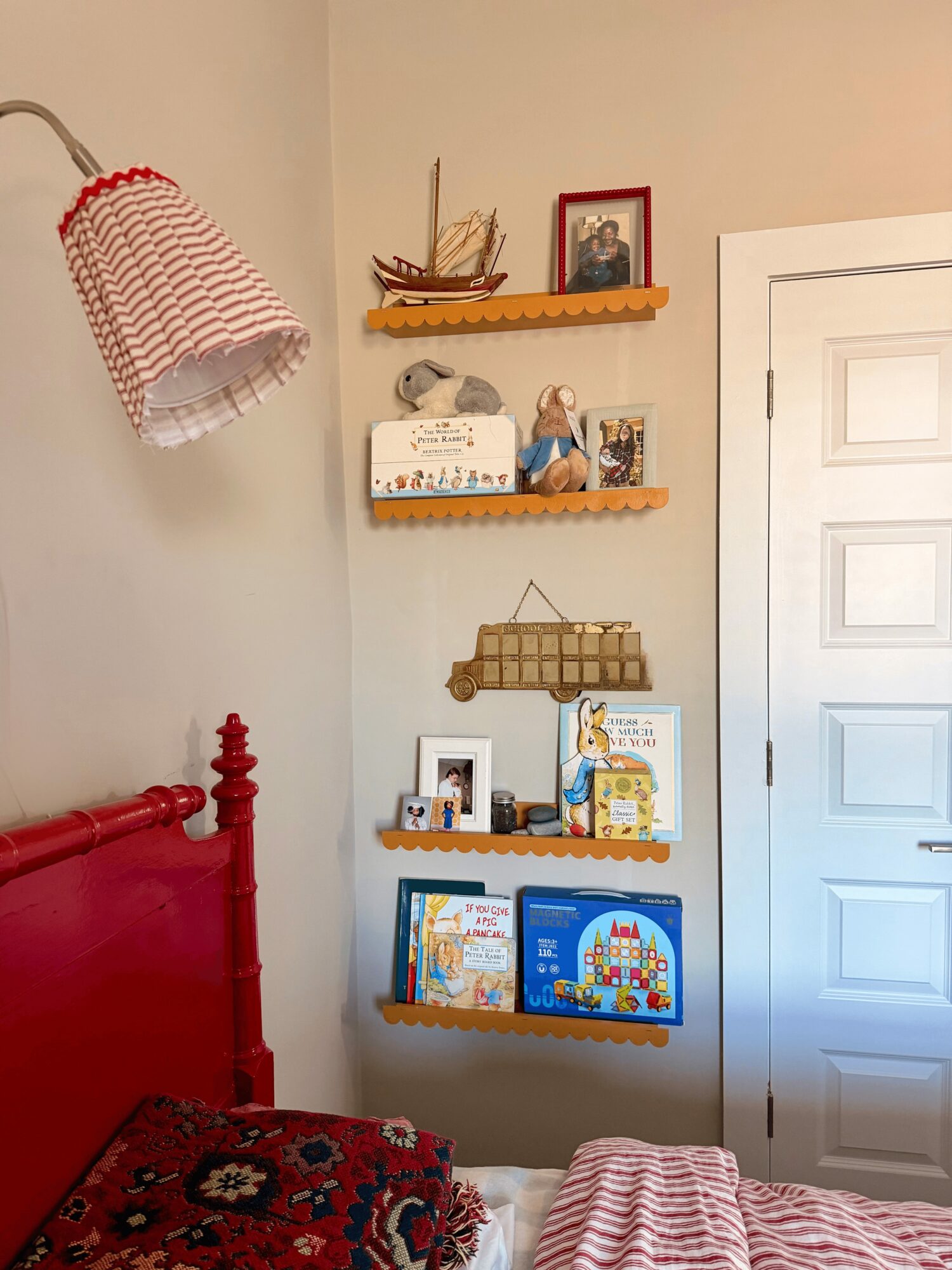 Corner of a room with a red bed, wall shelves holding books and decorations, and a white door.