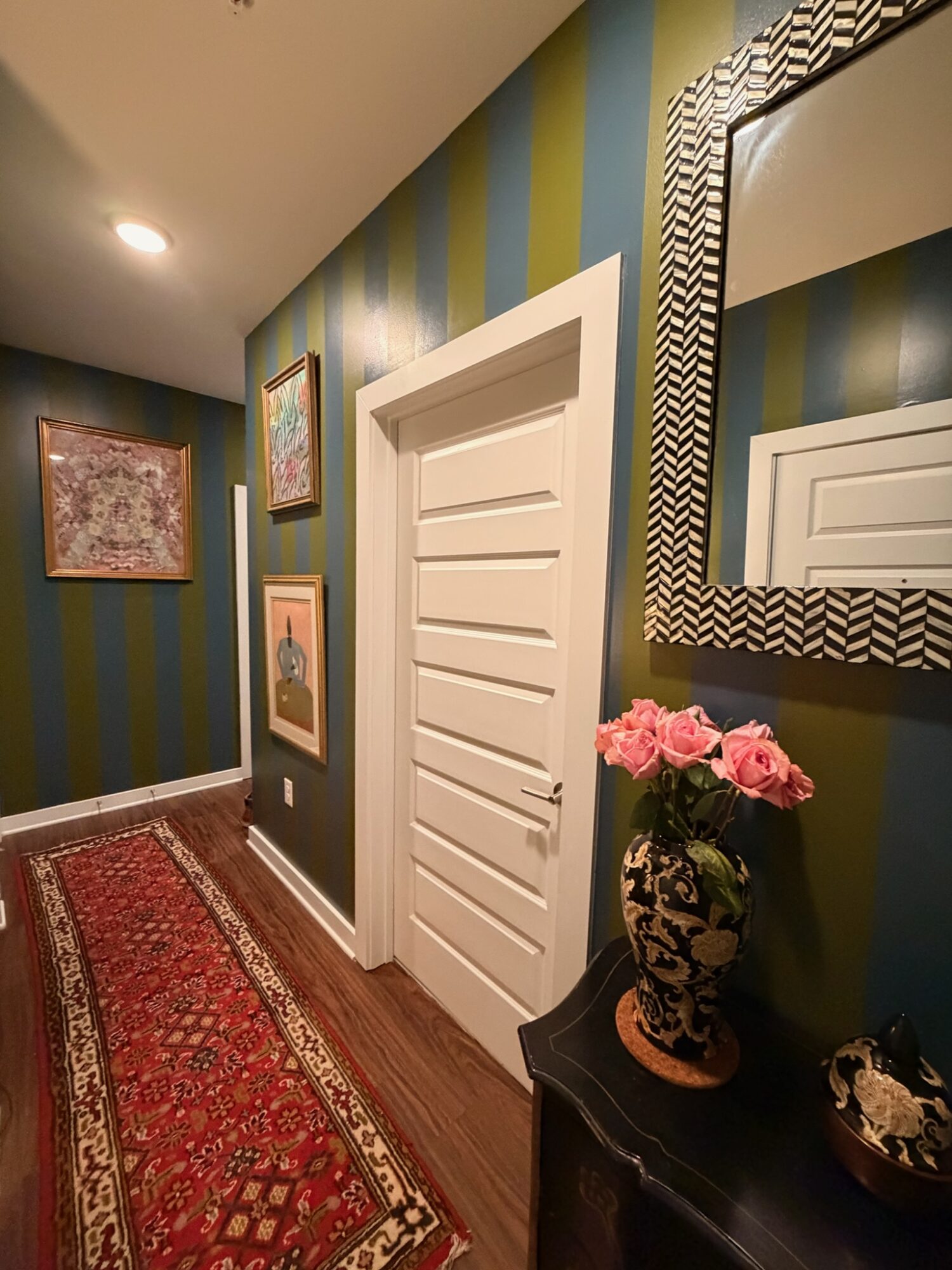 Hallway with striped green and blue walls, framed artwork, a white door, a mirror, and a vase with pink roses on a black table.