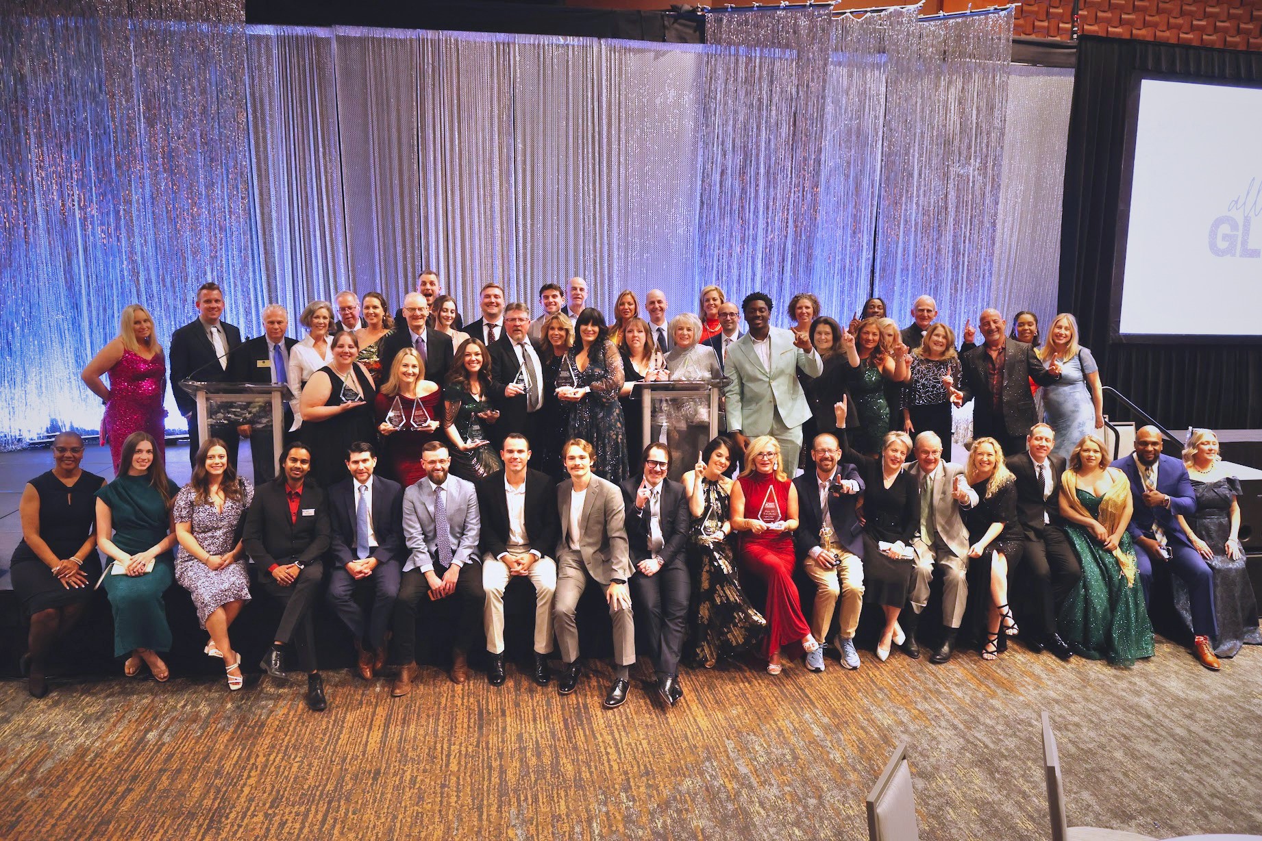 Group of people on stage at an event, with a blue curtain backdrop and a screen on the right.
