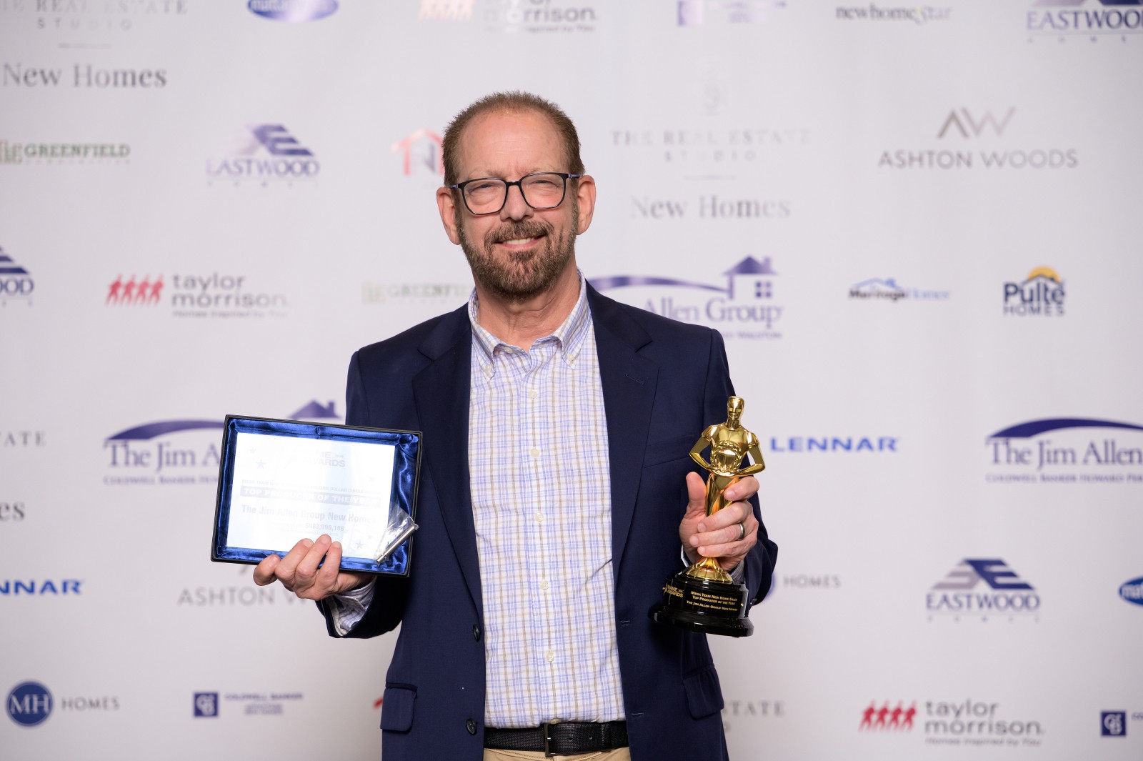 Man with glasses and beard holding a framed certificate and a trophy, smiling, in front of a step-and-repeat backdrop.