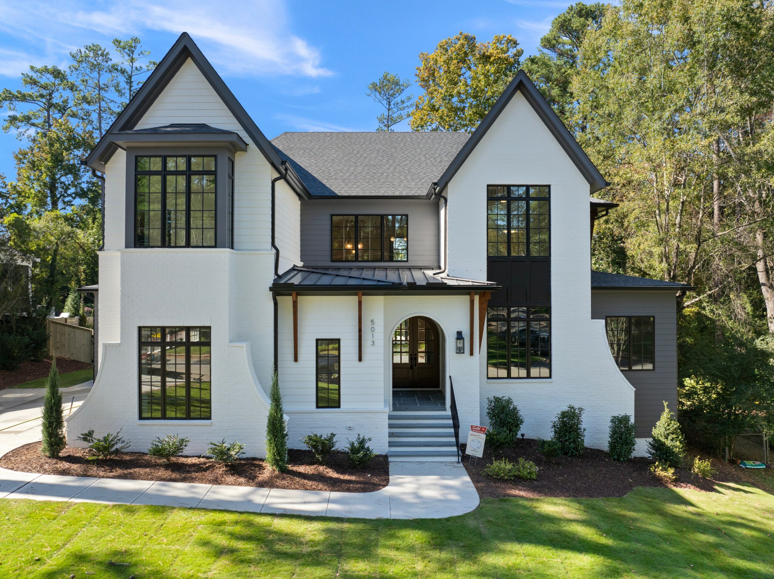 Two-story white house with black window frames, front steps, and a landscaped yard, surrounded by trees under a blue sky.