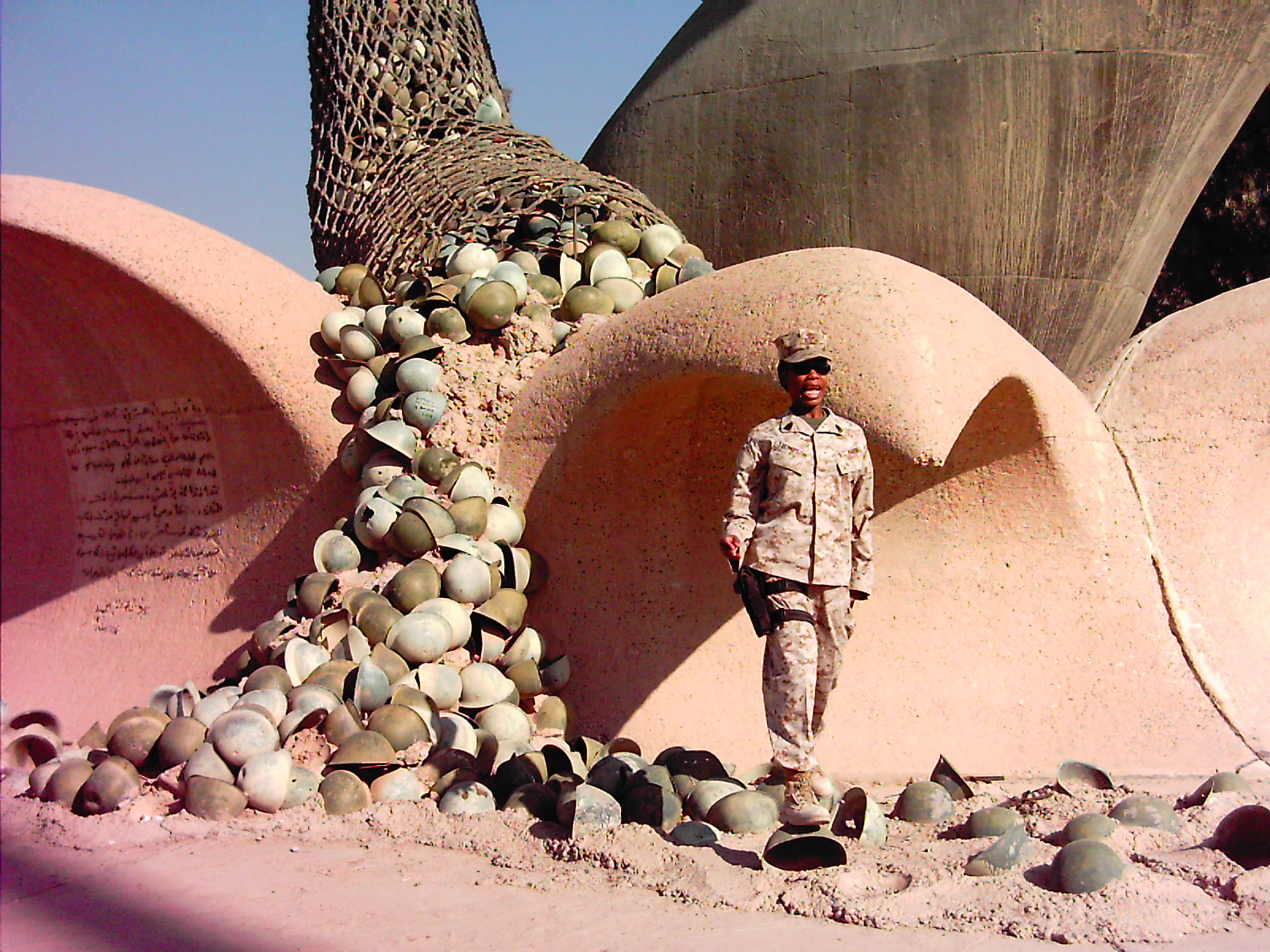 A soldier in uniform stands next to a large sculpture with a tree and numerous spherical objects, against a clear sky.