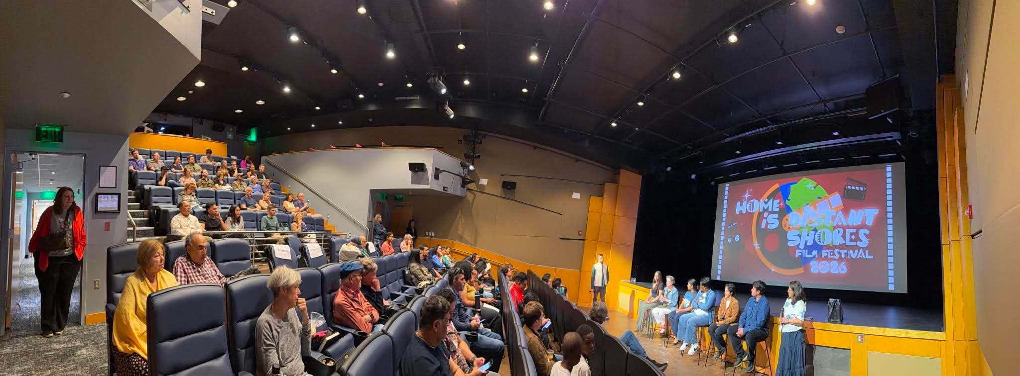Auditorium with people seated, a stage with a large screen, and a speaker at the front.