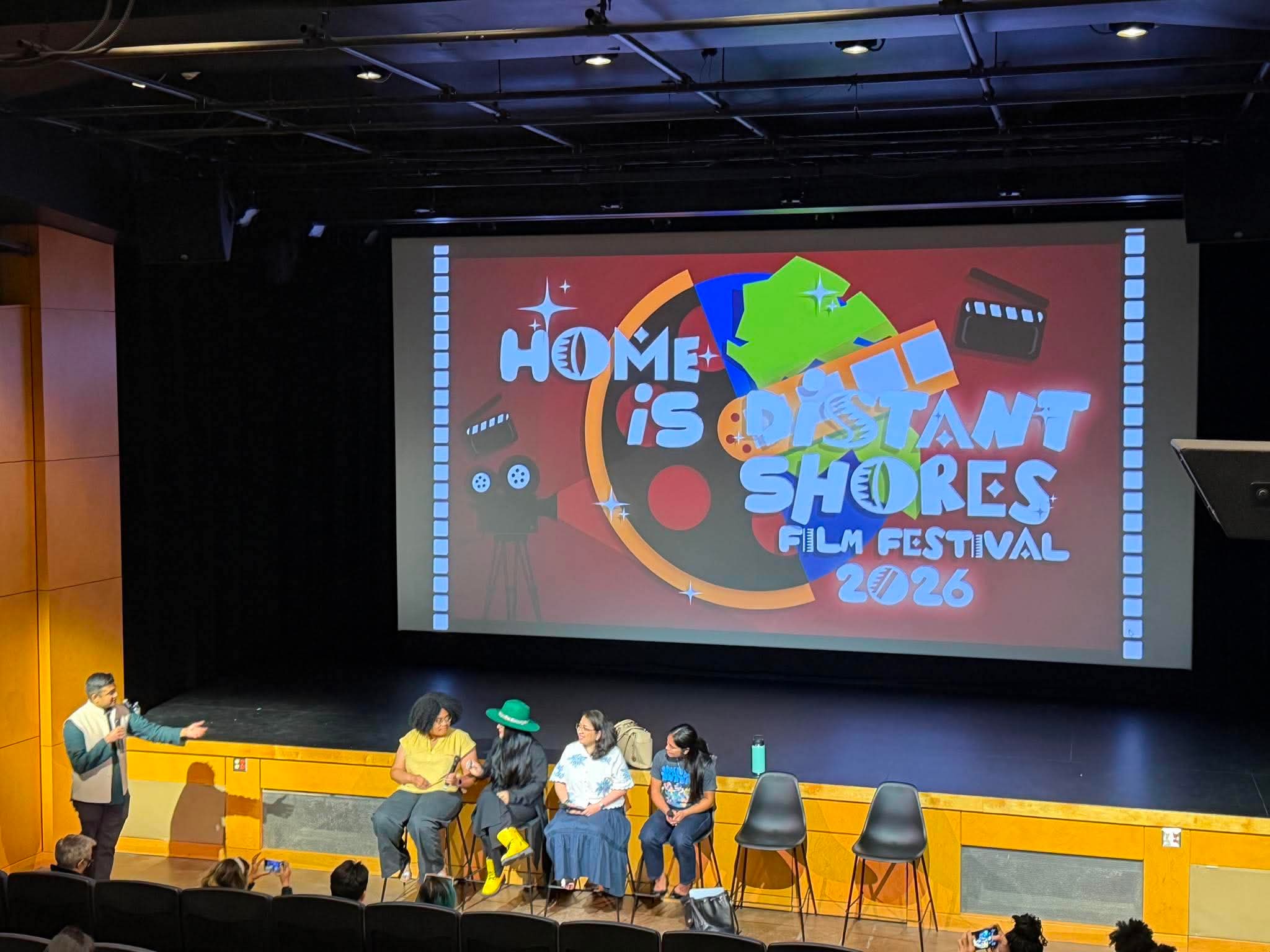 People sitting on chairs on stage during a panel discussion at a film festival, with a large colorful screen behind them.