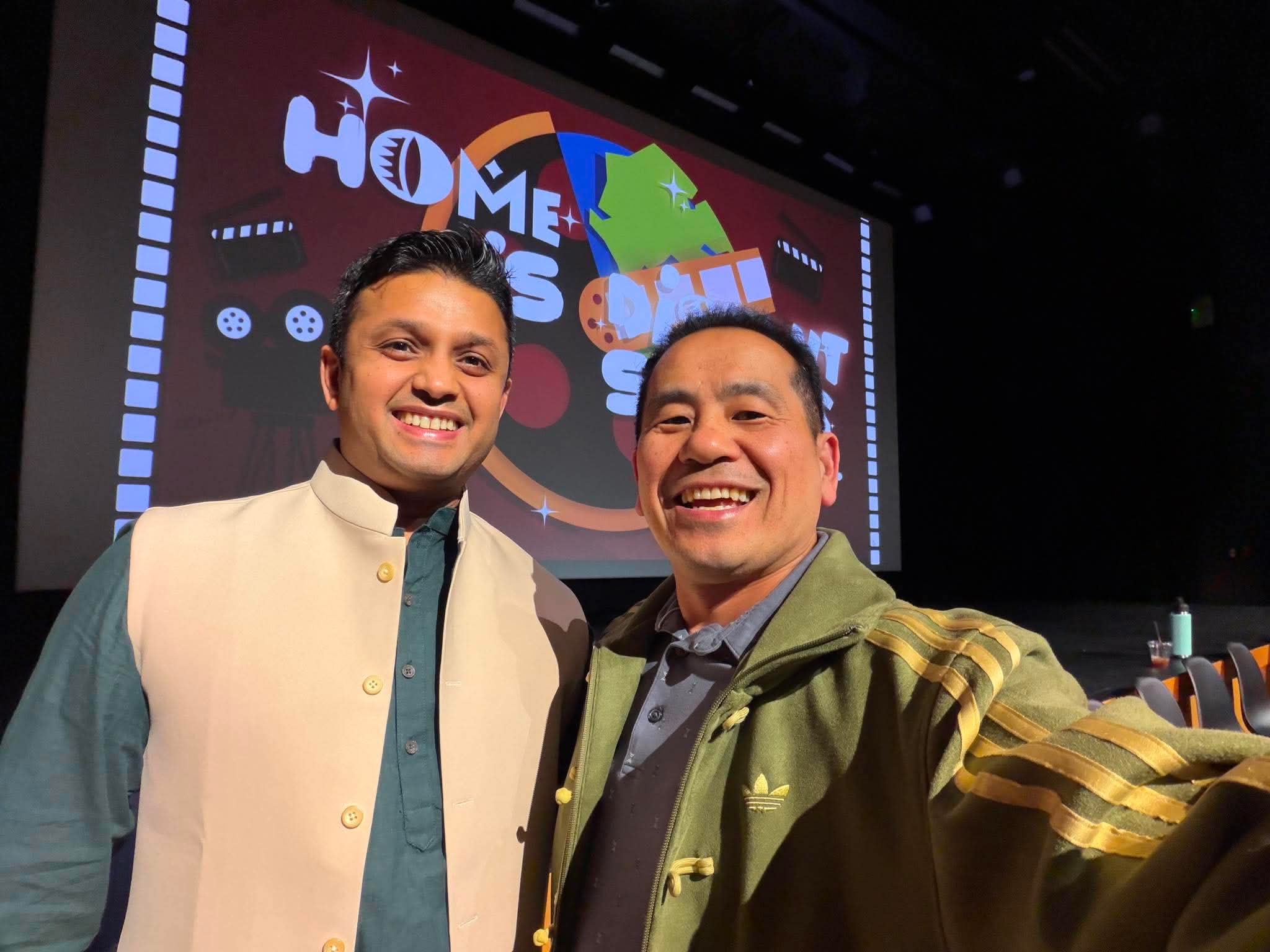 Two smiling men taking a selfie in front of a screen with a colorful logo that says 'HOMES'.