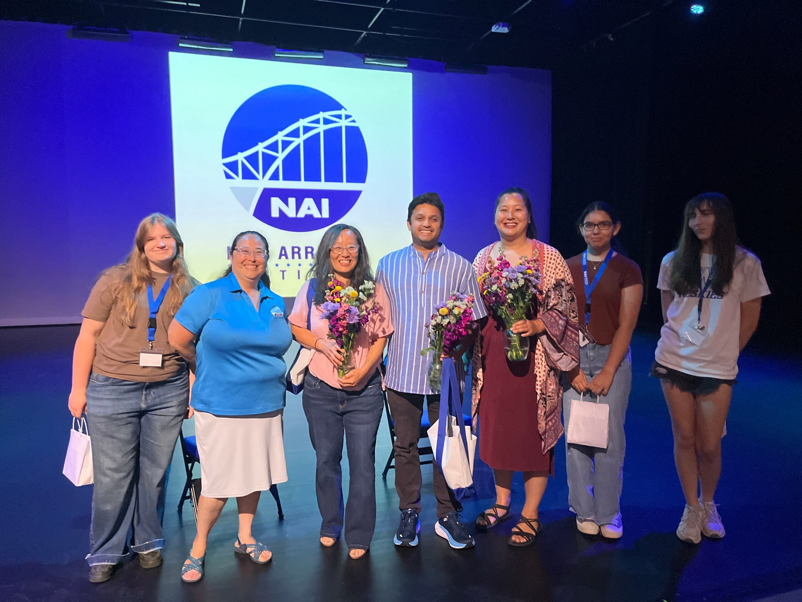 Group of seven people standing on stage with a large screen displaying a logo behind them, some holding flowers.