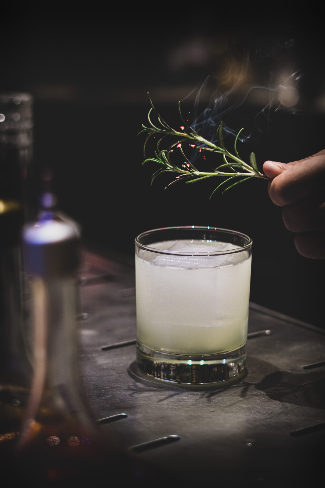 Hand holding a sprig of herbs above a cloudy drink in a glass on a dark surface.