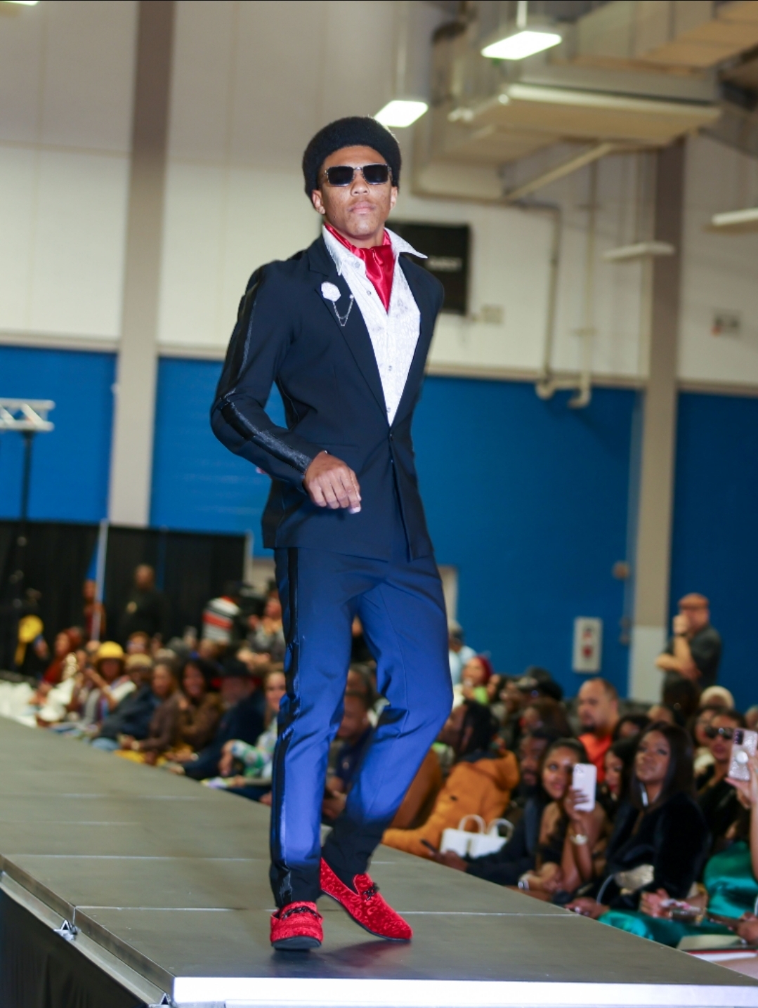 Male model walking on runway wearing sunglasses, black jacket, white shirt, blue pants, red shoes, in indoor event with audience