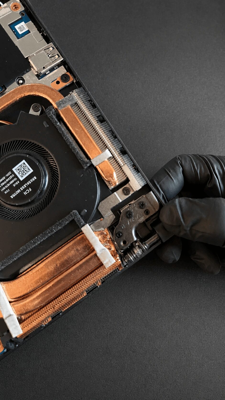 Person wearing a black glove holding a partially disassembled electronic device with a cooling fan and copper heat sink, on a black surface.