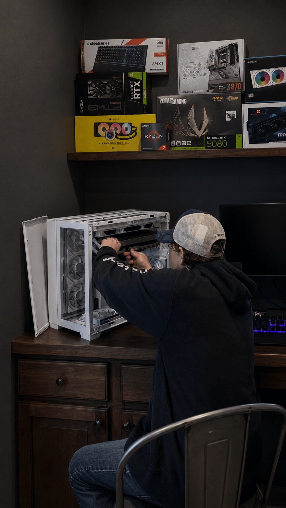 Person wearing a white cap and black hoodie working on a computer inside a store, with shelves of electronics above.