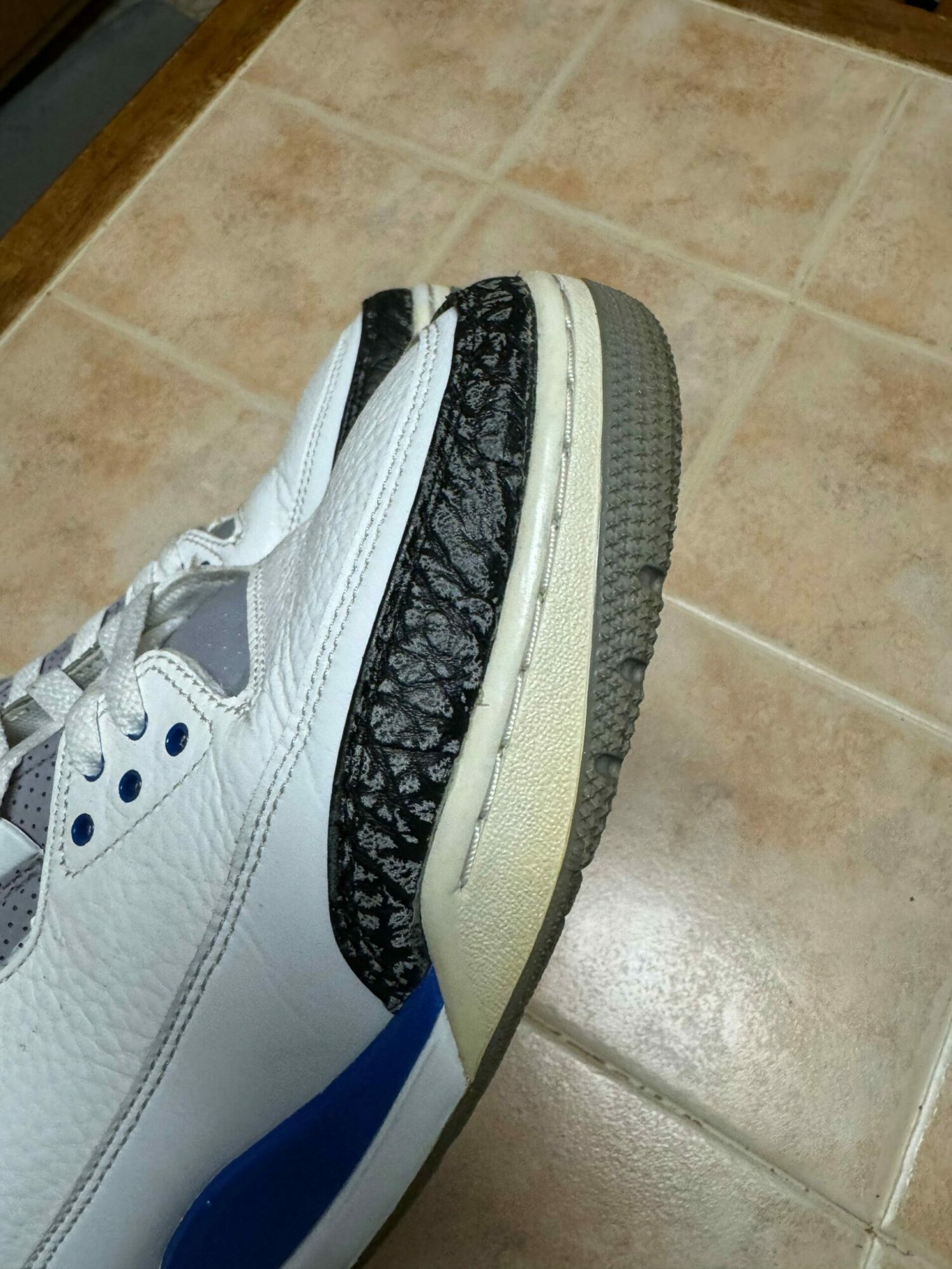 Close-up of a white sneaker with black and blue accents on a tiled floor.