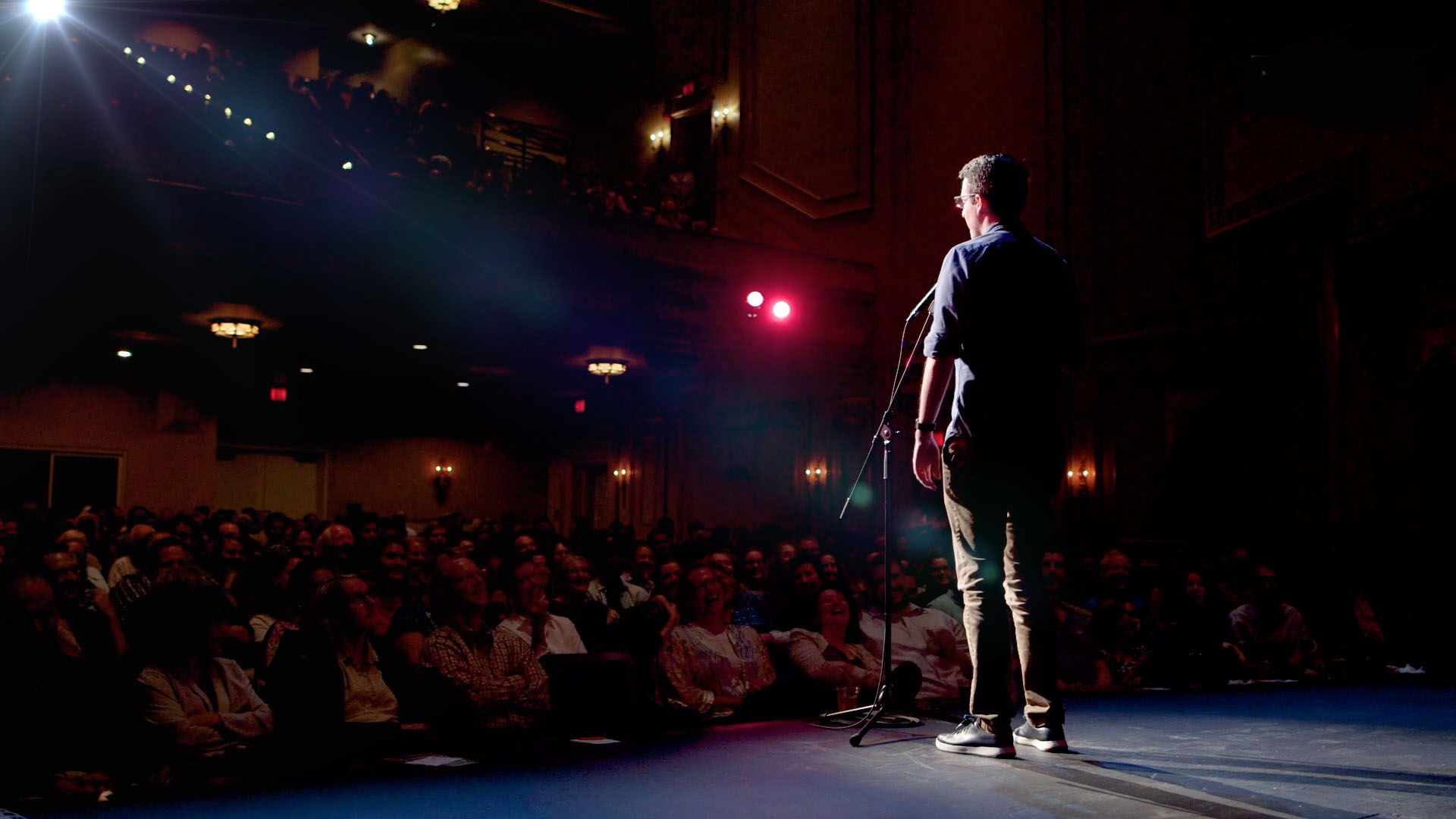 Performer stands on stage facing audience in a dark theater with colorful stage lights, audience seated in front.