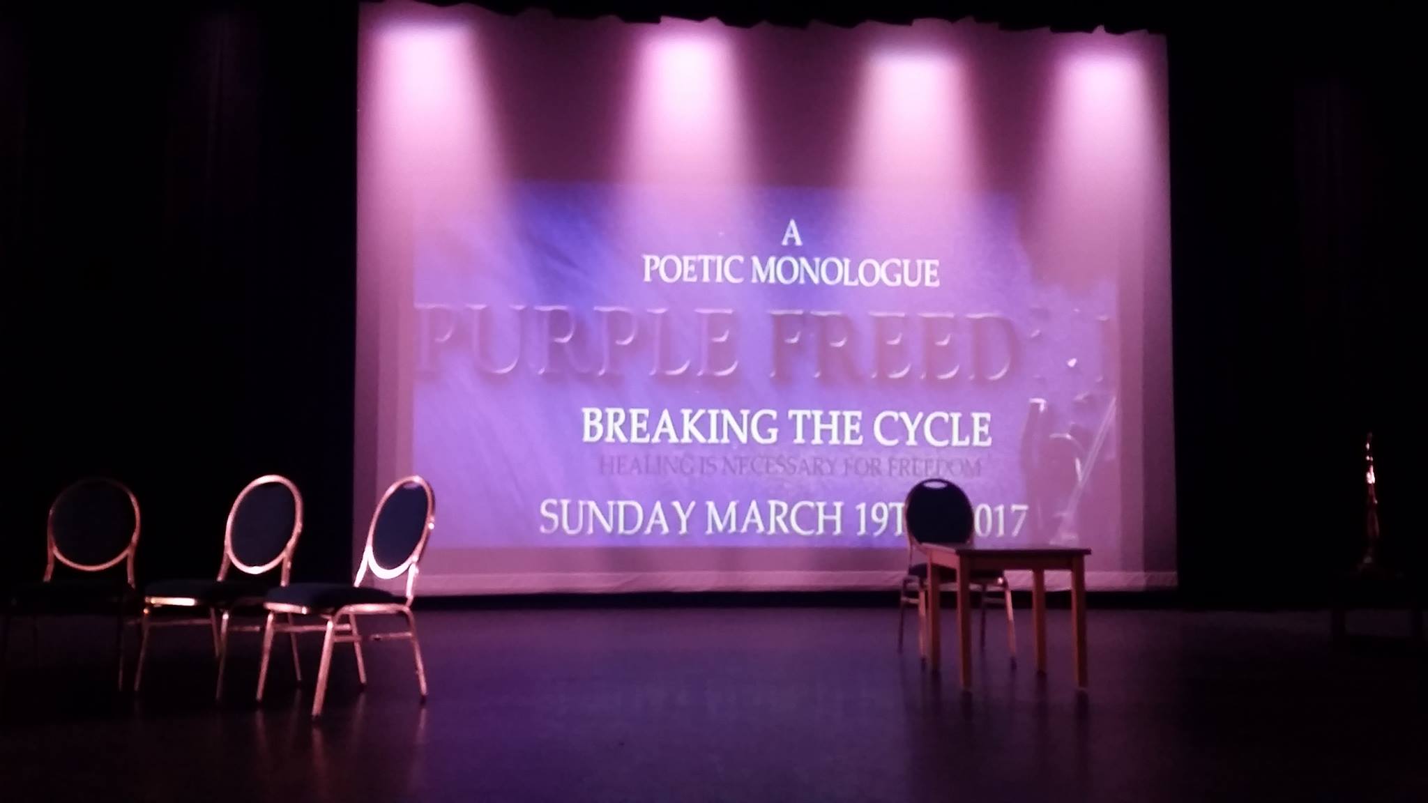 Stage with chairs and a table, large screen displaying event details, purple lighting, and curtains in the background.