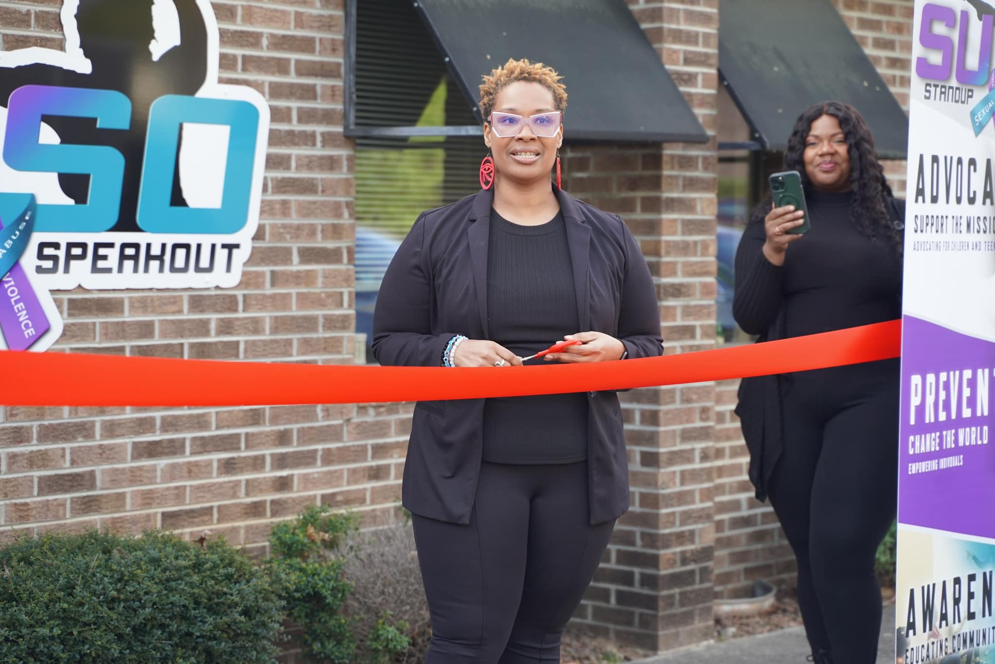 Two women standing outside a building during a ribbon-cutting event, one smiling and holding a phone, the other using a phone.