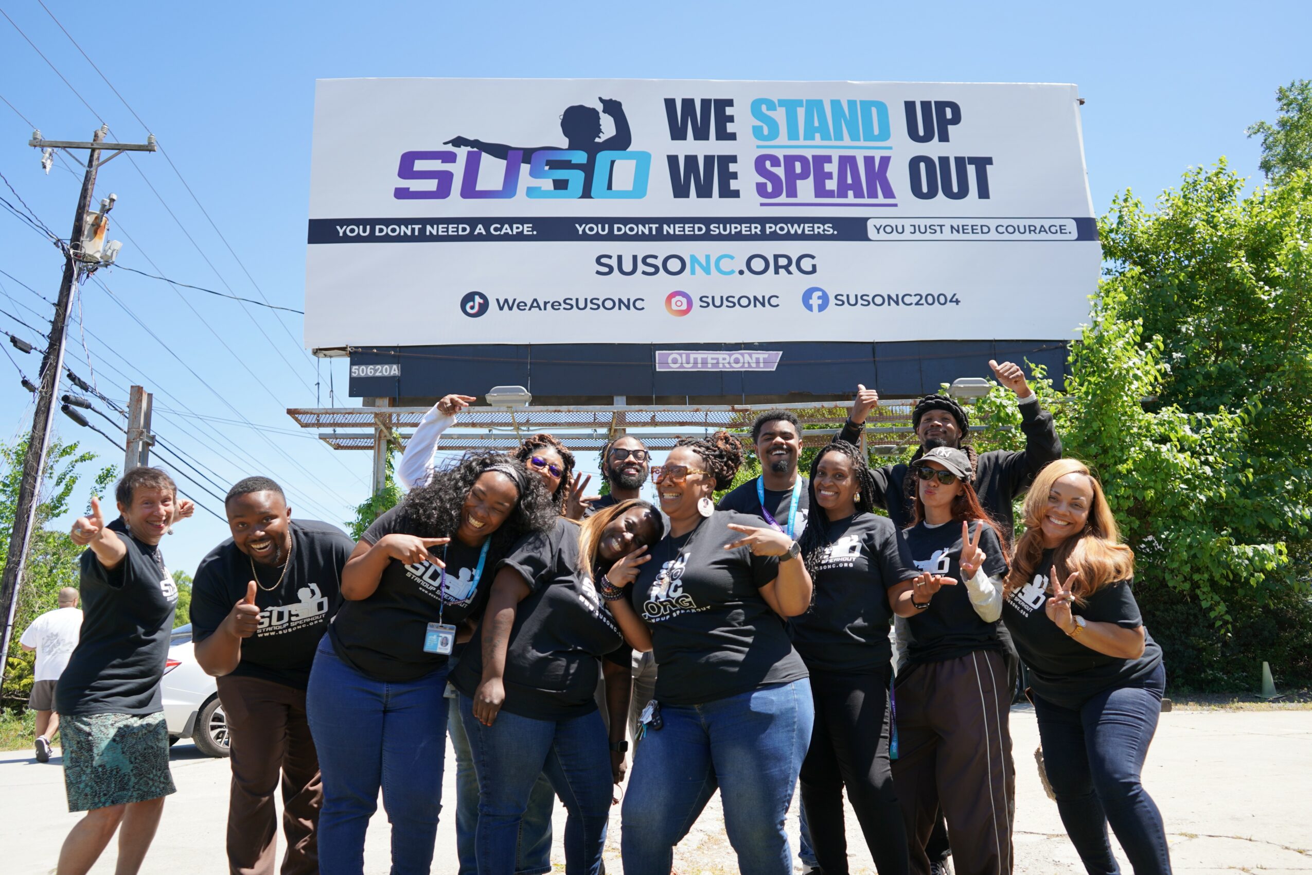 Group of people standing outdoors in front of a large banner supporting standing up and speaking out, smiling and holding fists up.