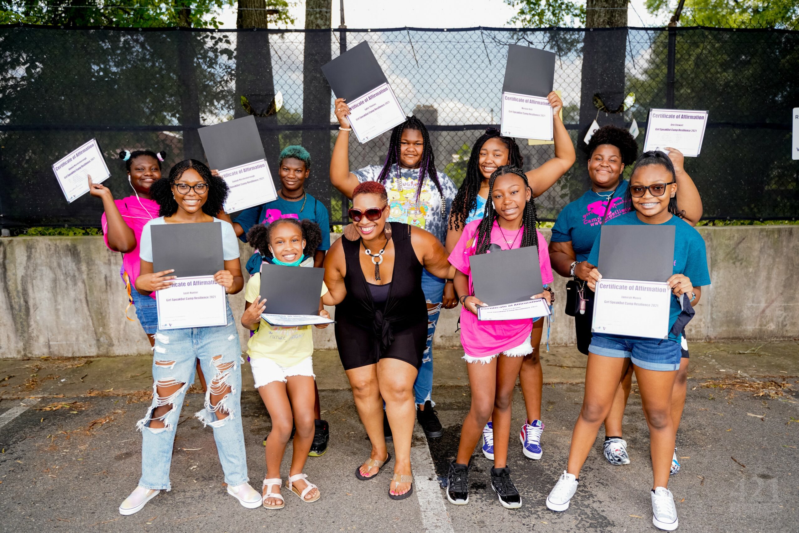 Group of children and an adult woman holding certificates and laptops outdoors on a tennis court, smiling.