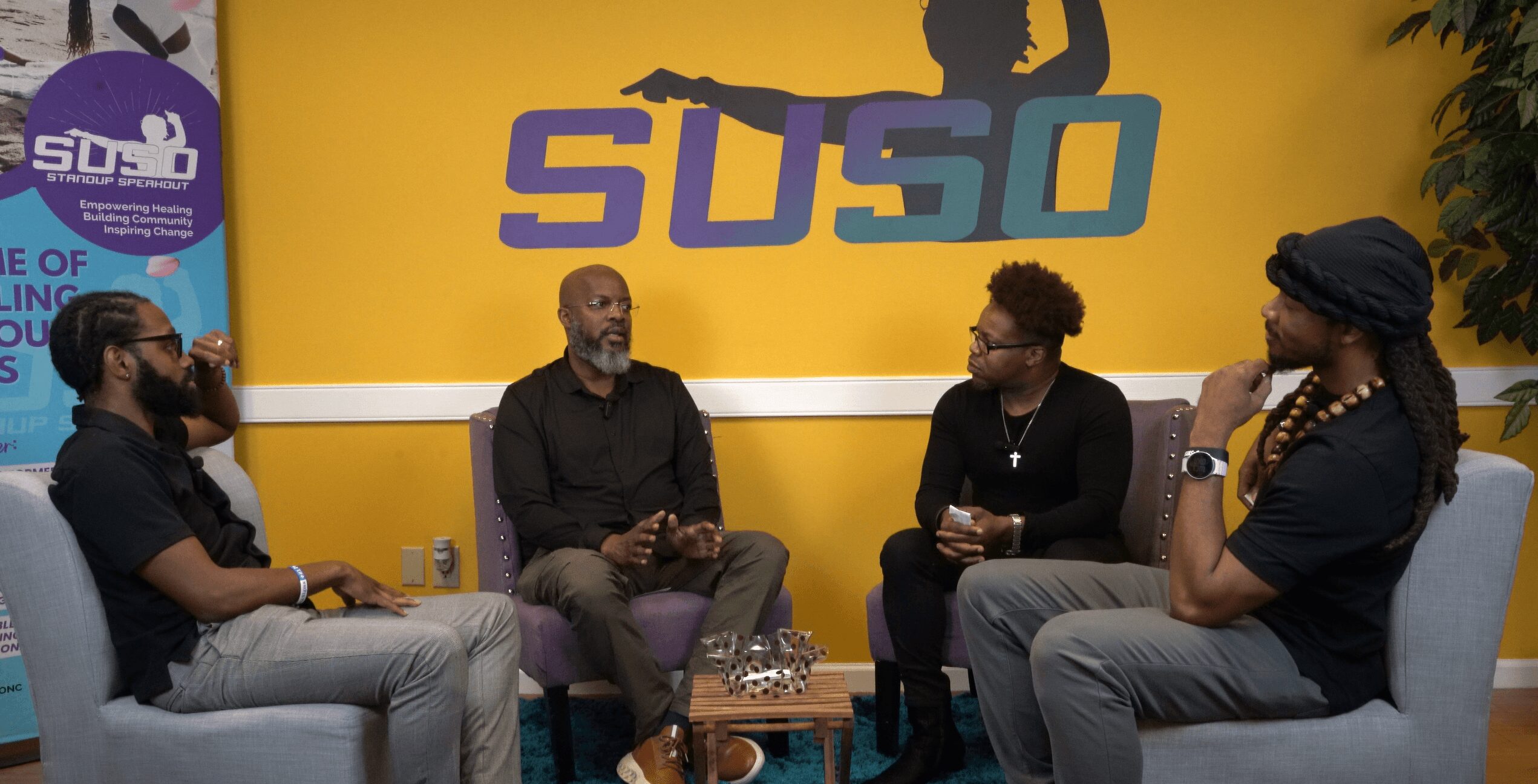 Four people sit on chairs in a room with a yellow wall and a large logo reading 'SUSO' behind them.
