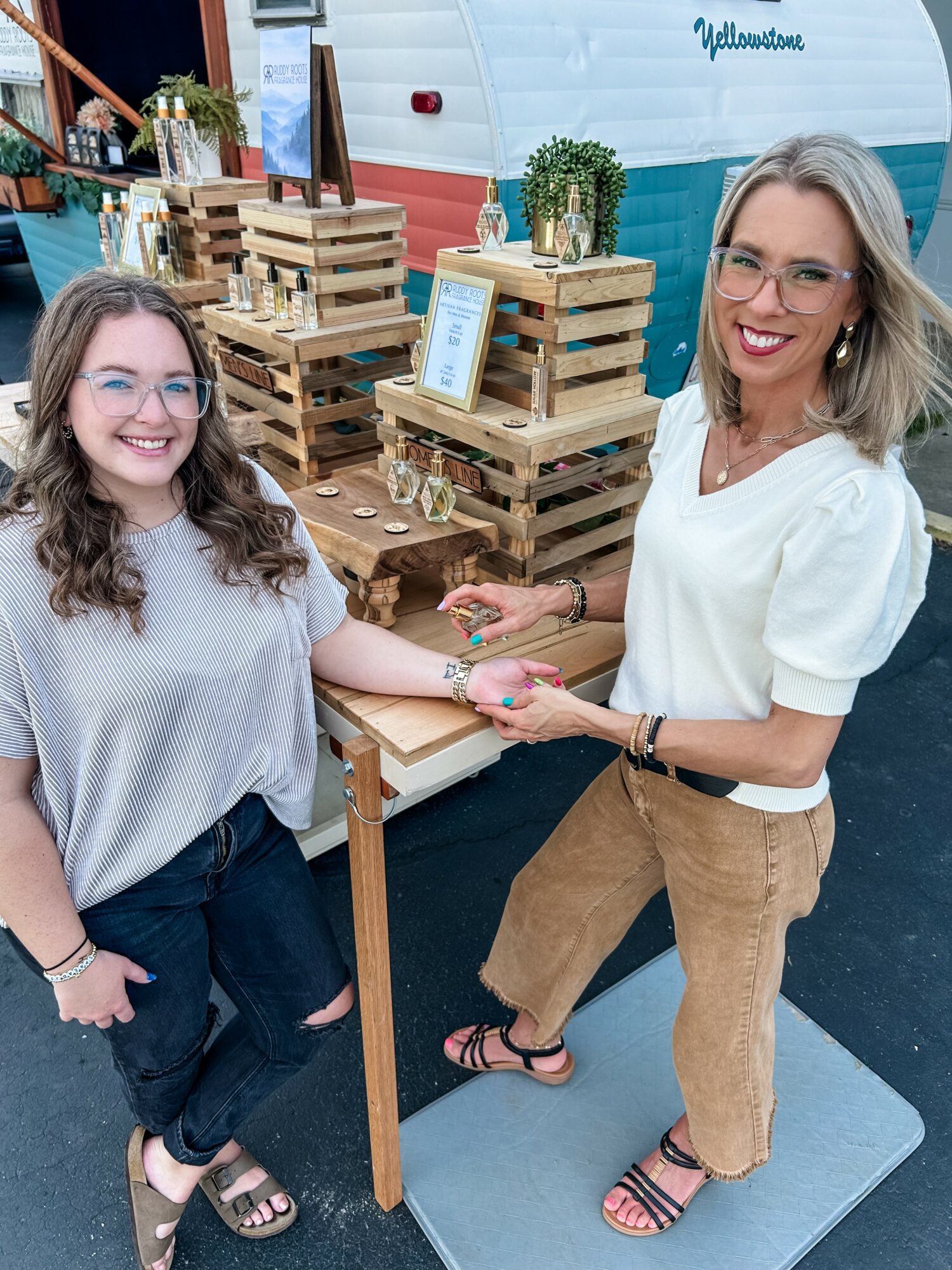 Two women smiling and holding hands at an outdoor event with wooden pallet displays and potted plants.