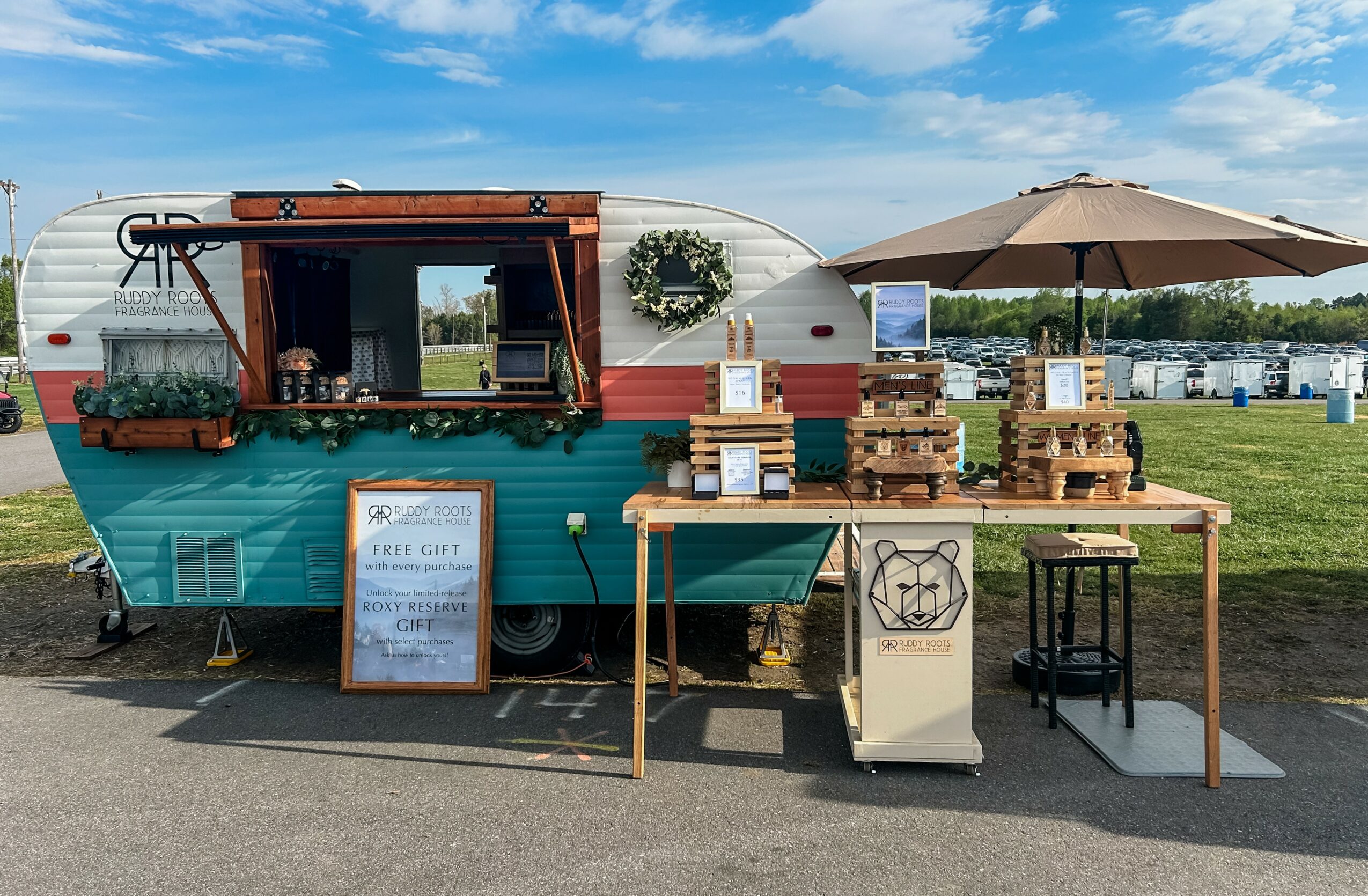 A vintage camper converted into a mobile shop with outdoor seating and display tables, set on a grassy area under a blue sky.
