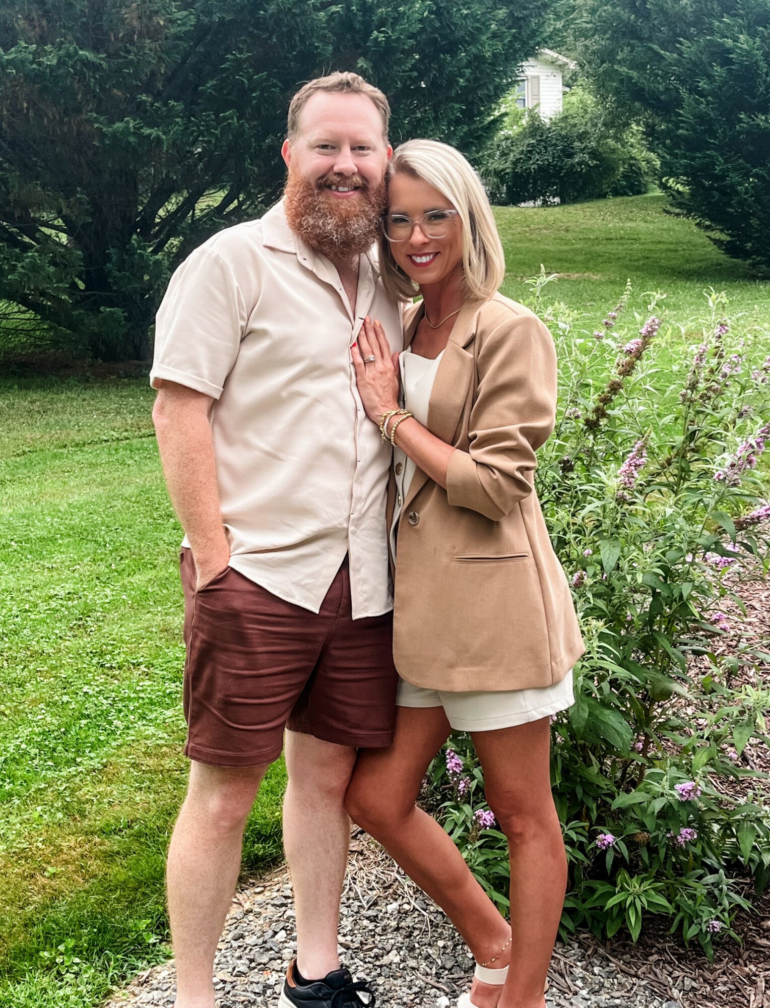 A man and woman stand outdoors on a grassy area with trees and flowers, smiling and embracing each other.