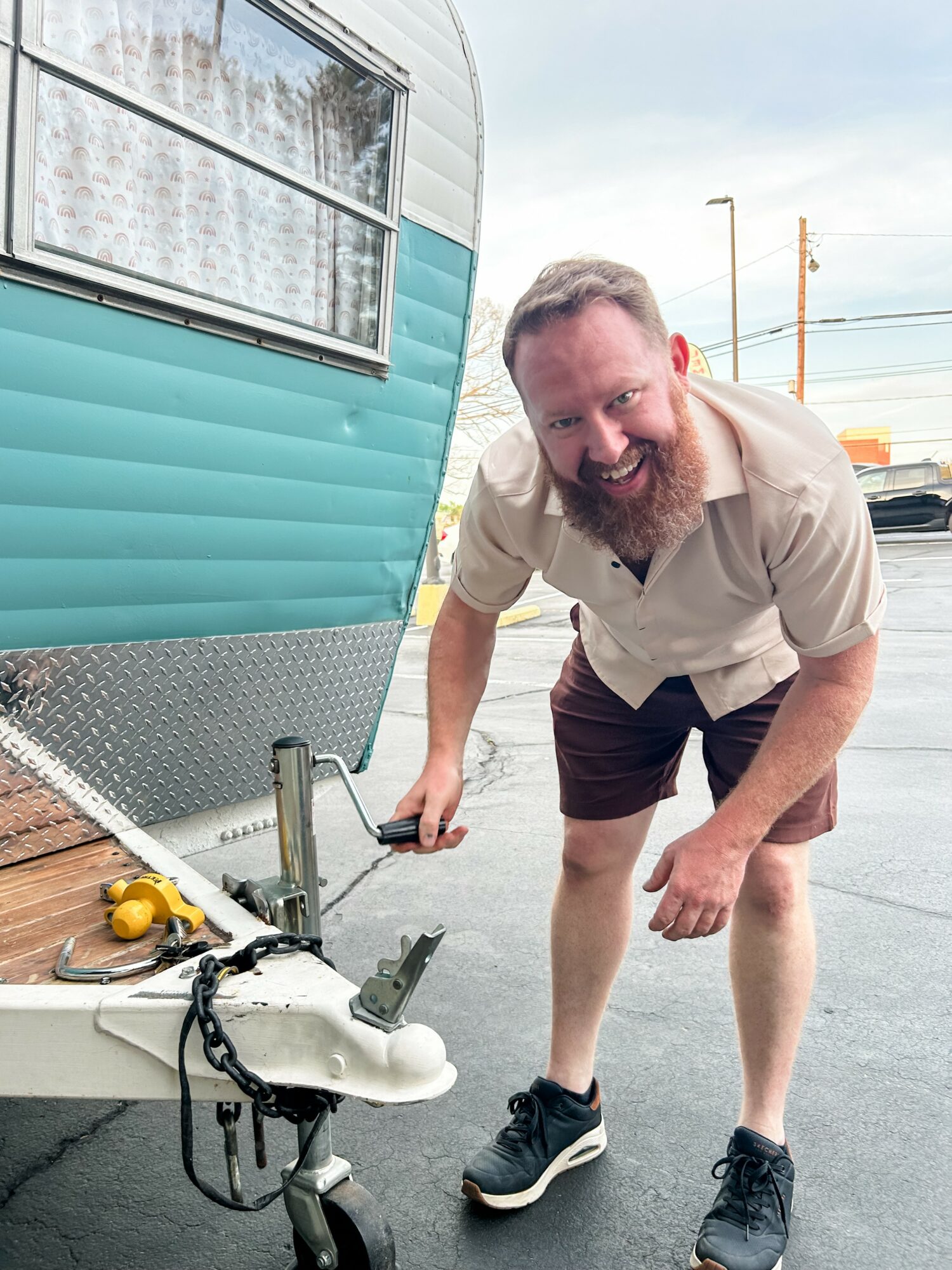 Man with beard smiling, bending over near a teal and white camper trailer, holding a tool, in an outdoor parking lot.