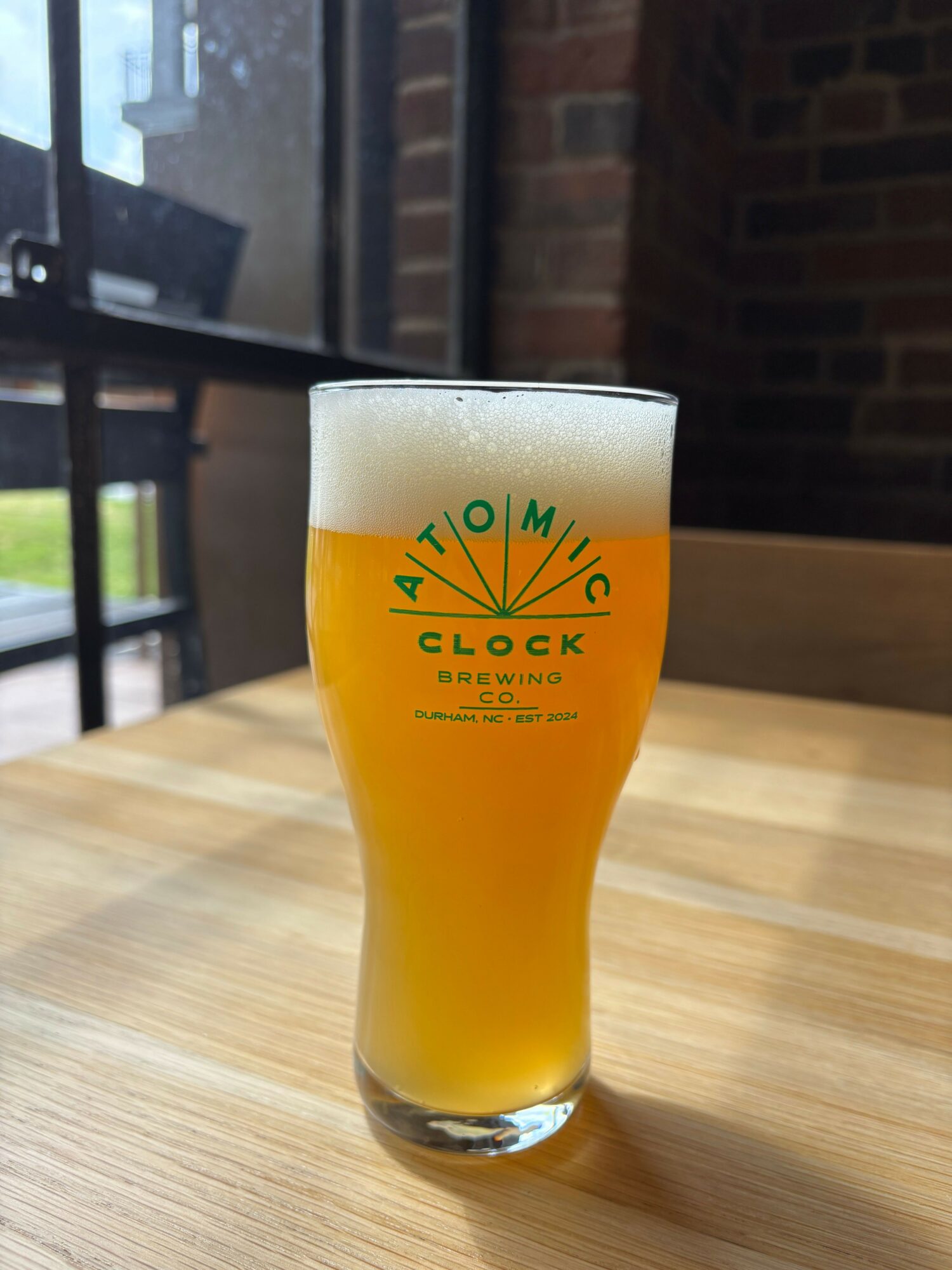 A glass of orange-colored beer with foam on top, placed on a wooden surface indoors.
