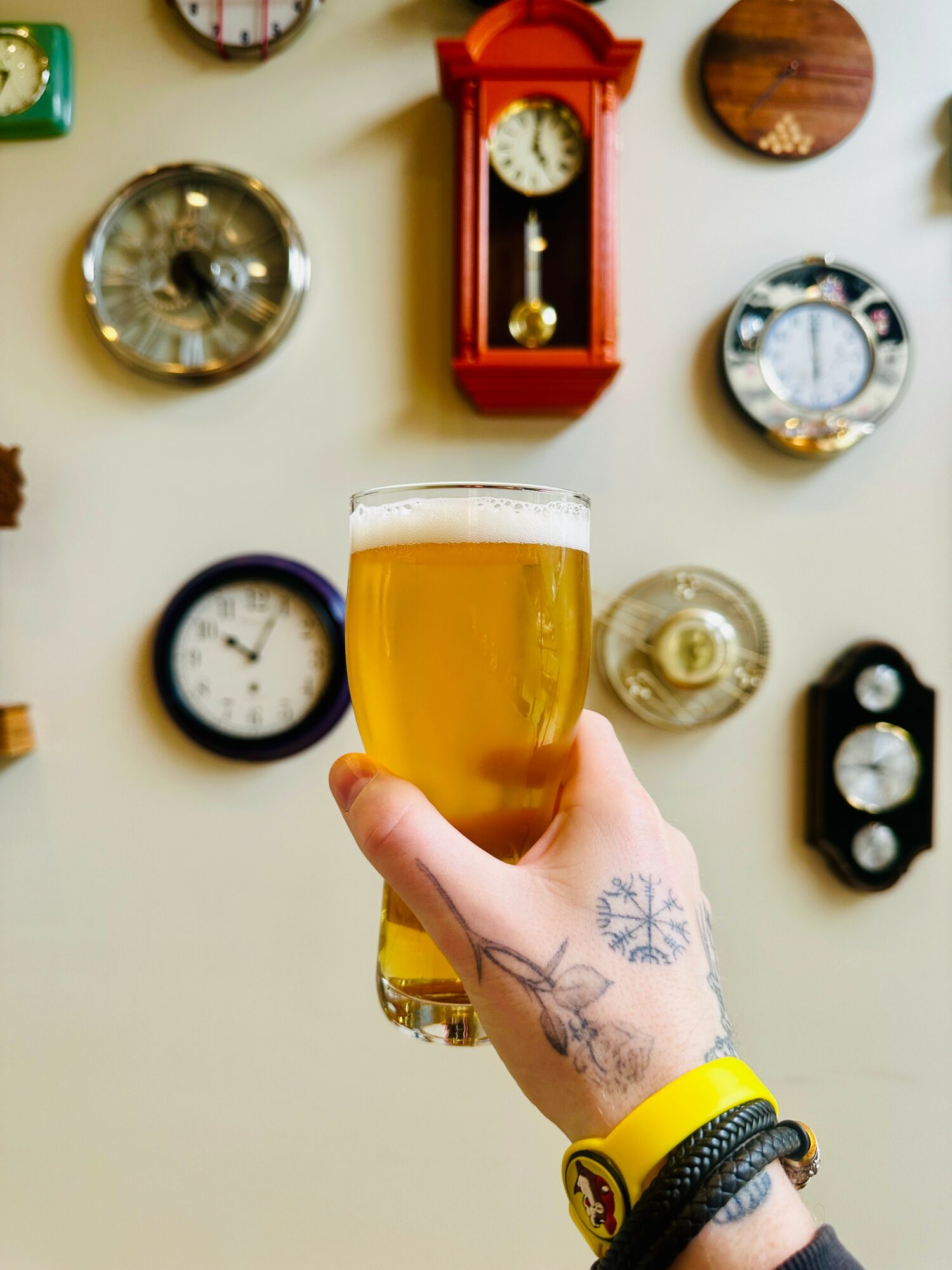Hand holding a glass of beer with a tattooed hand and wristbands, wall with various clocks in the background.