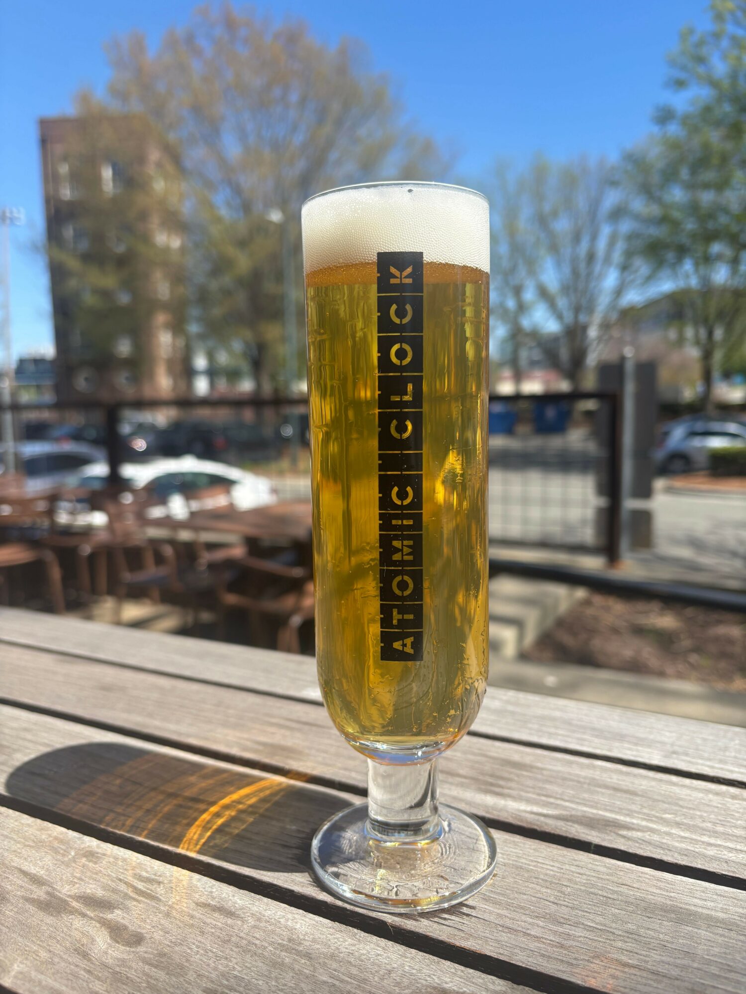 A glass of beer with foam on top, placed on a wooden table outdoors, with trees and buildings in the background.