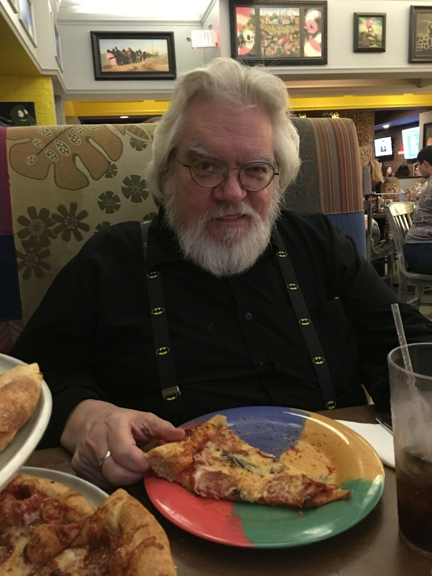 Older man with glasses and white beard sitting at a table with pizza, smiling, in a restaurant setting.