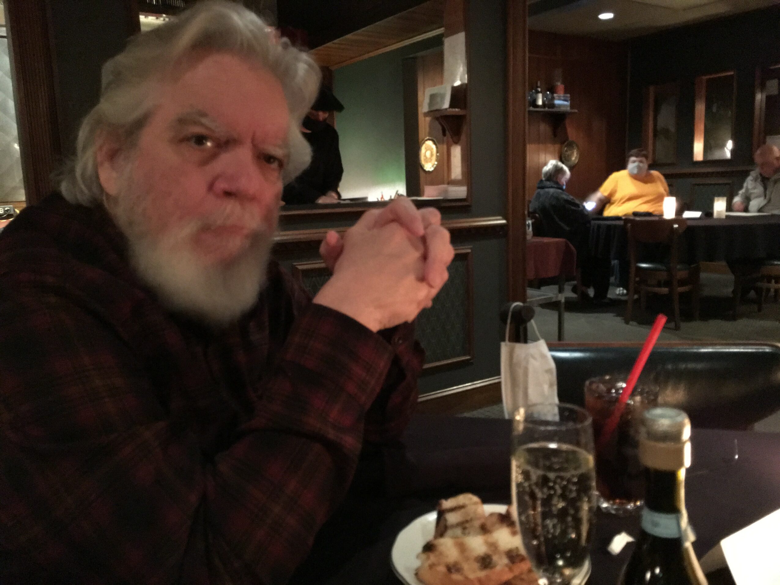 Older man with gray hair and beard sitting at a restaurant table, holding a fork, with drinks and food in front.