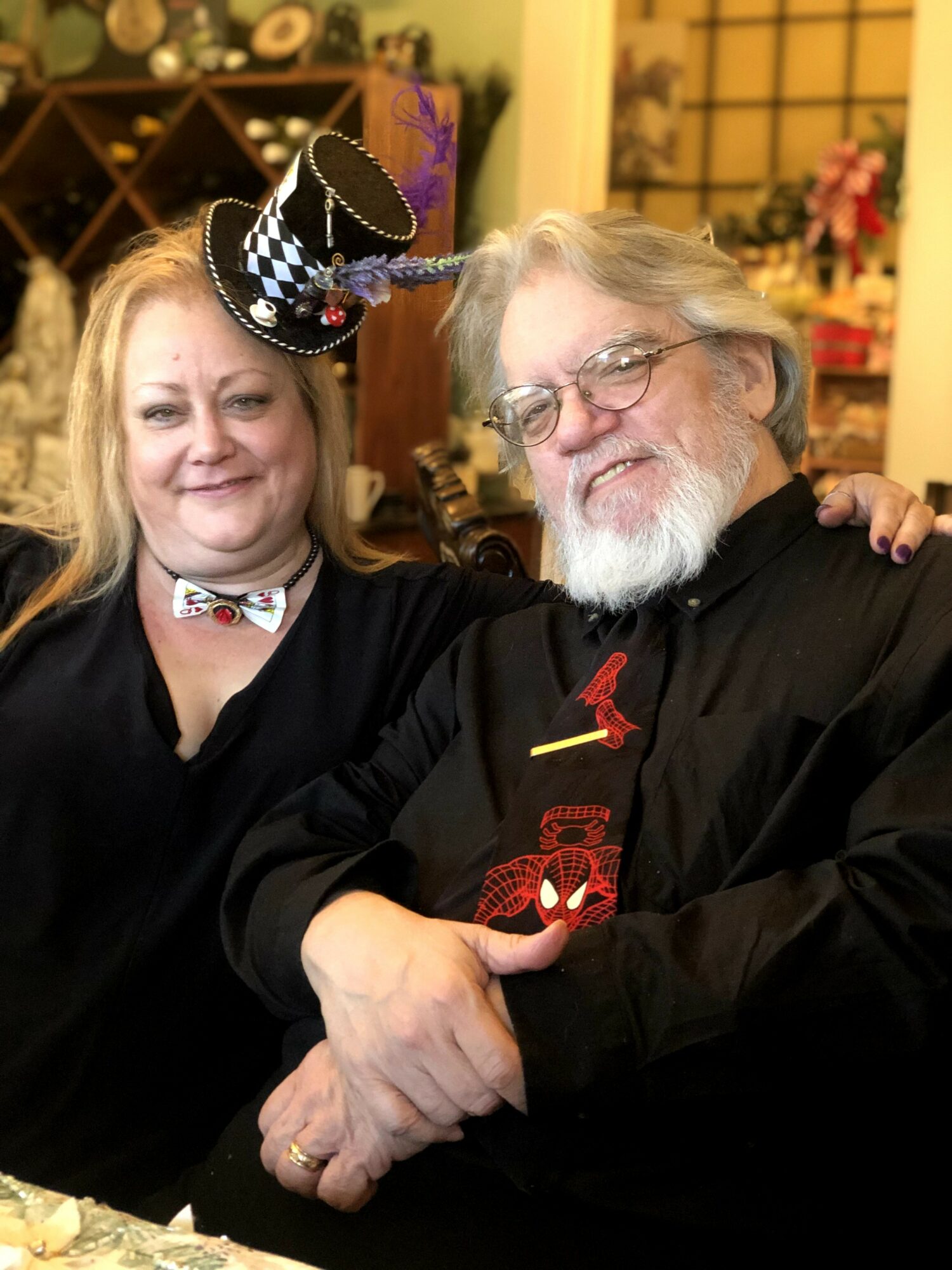 Two people smiling, one wearing a festive hat and bow tie, the other with glasses and a beard, in a decorated room.