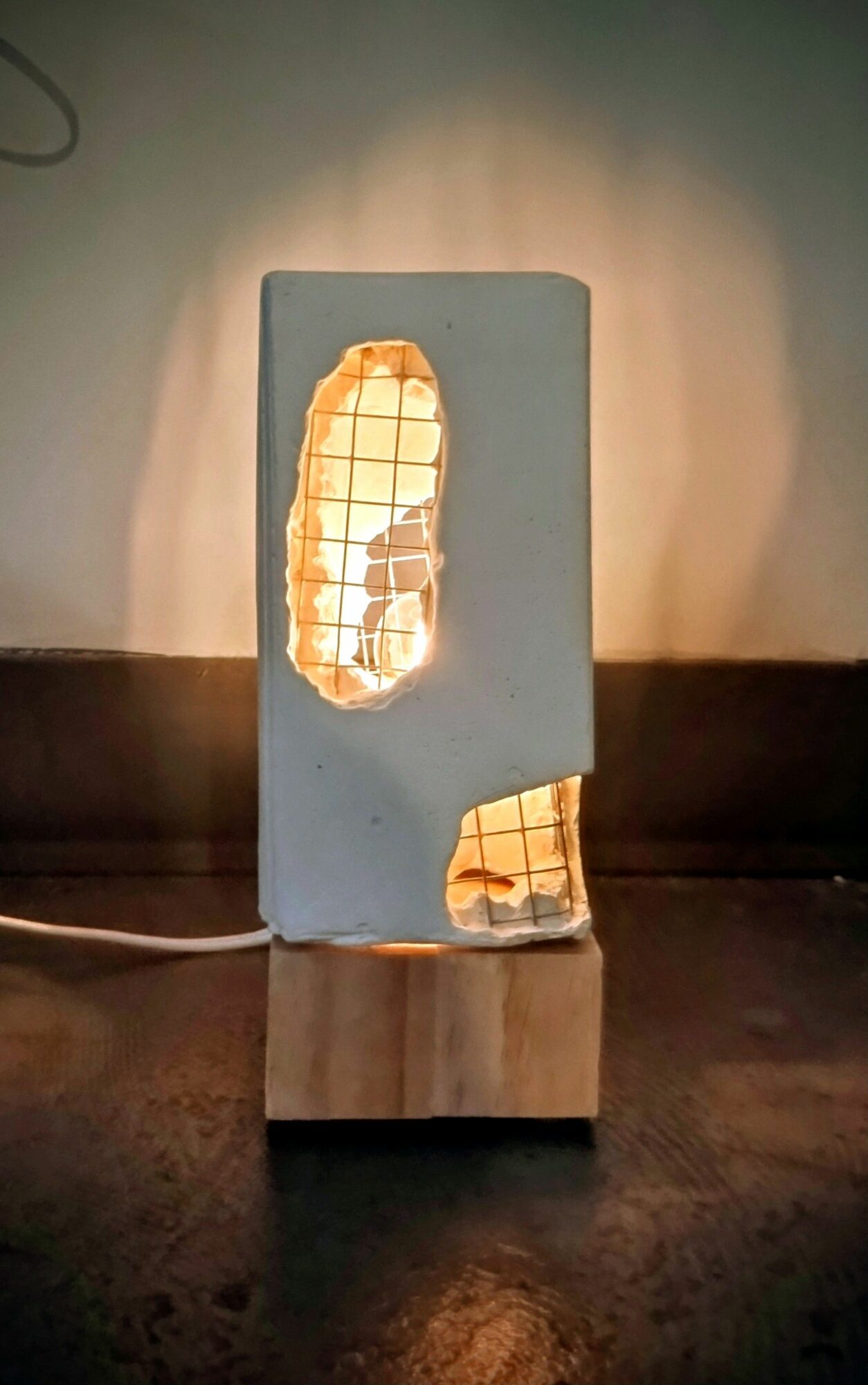 Lamp with a rectangular cutout and grid pattern, emitting warm light, on a dark surface with a wall background.