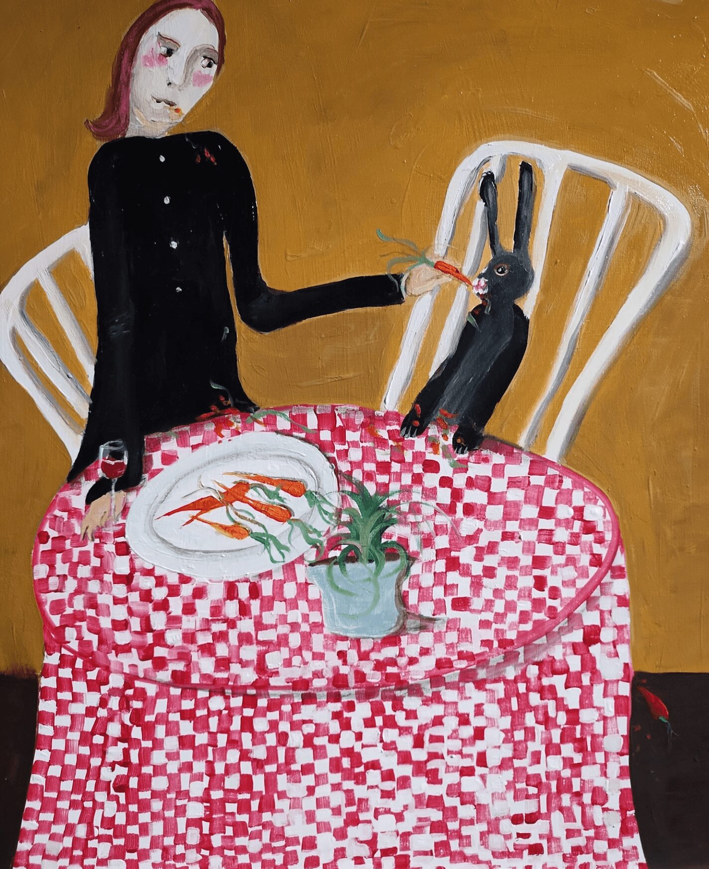 Woman with long black sleeves sitting at a table with a black rabbit, a plate with carrots, and a potted plant.