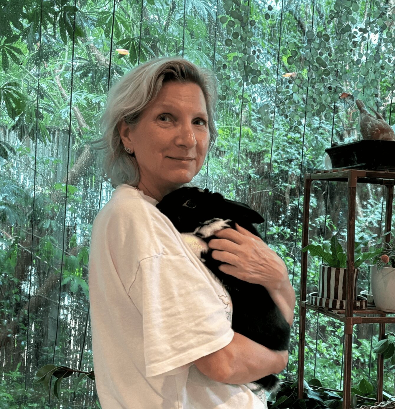 Woman with short blonde hair holding a black and white puppy in front of a lush green background.