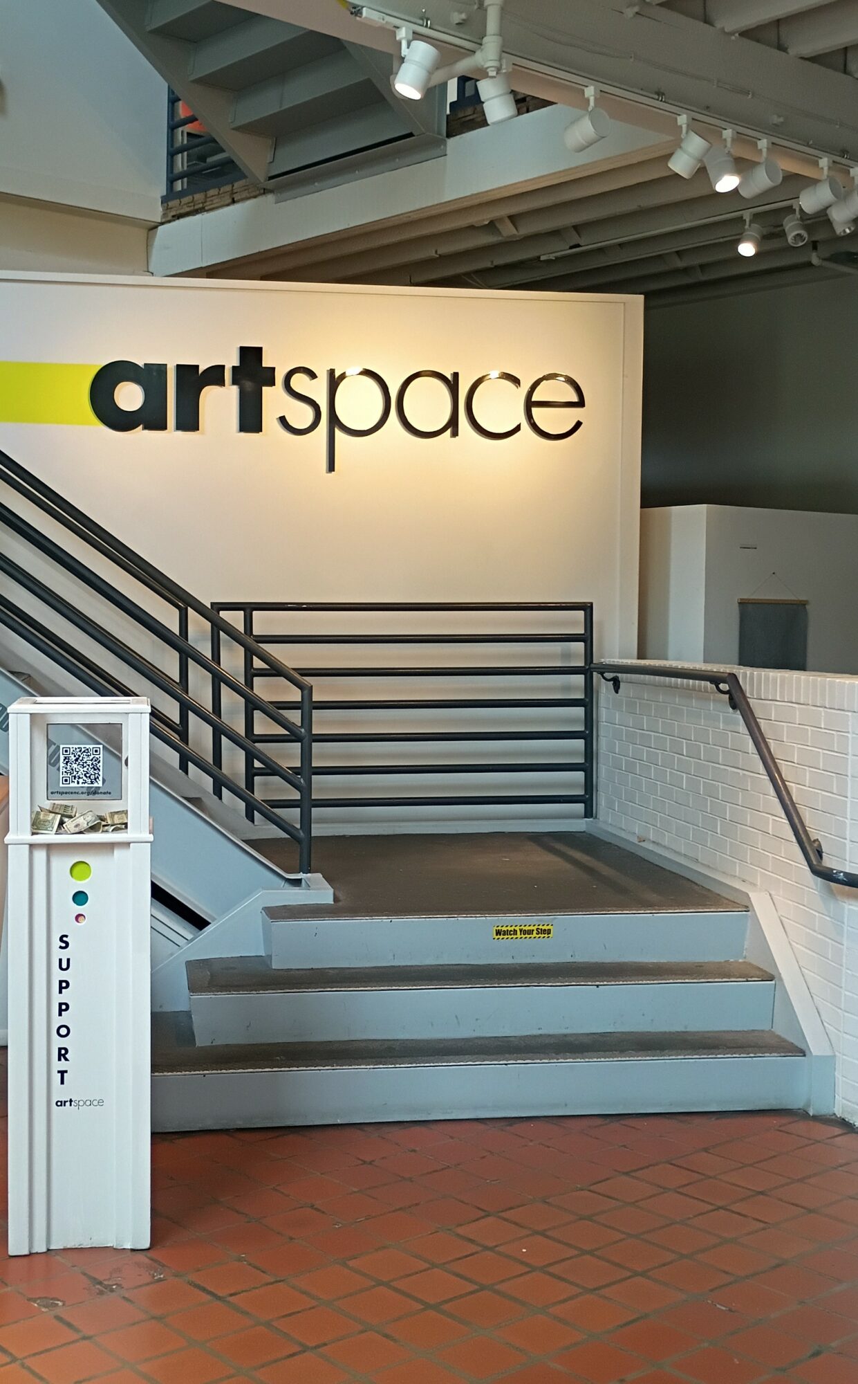 Sign reading 'art space' on a wall, staircase with black railing, and a support kiosk nearby, in an indoor setting.