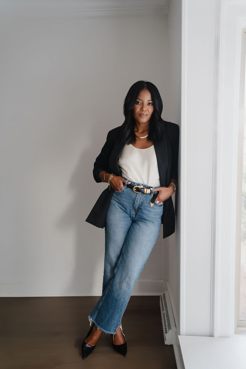 Woman standing indoors near a window, wearing a black blazer, white top, blue jeans, and black heels.