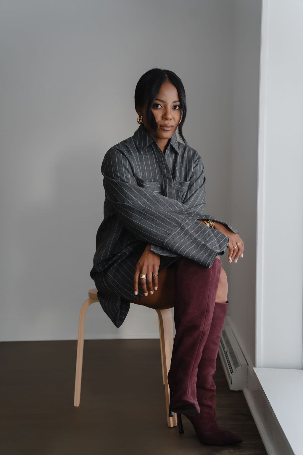 Woman sitting on a chair, wearing a dark striped shirt and maroon pants, in a room with plain walls and a window.
