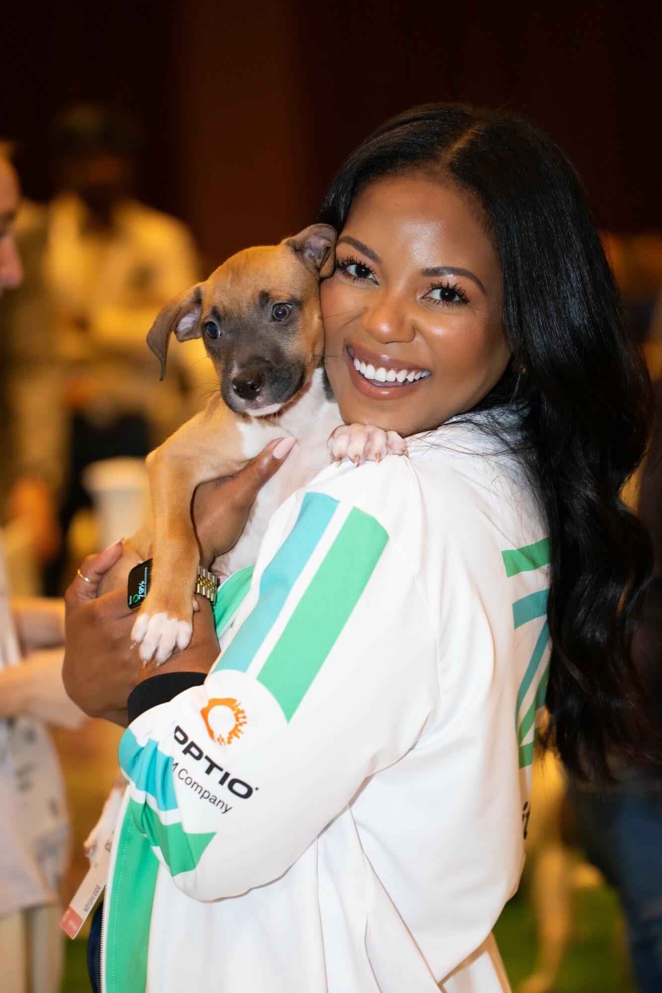 Smiling woman holding a small puppy, indoor setting with blurred background of people.