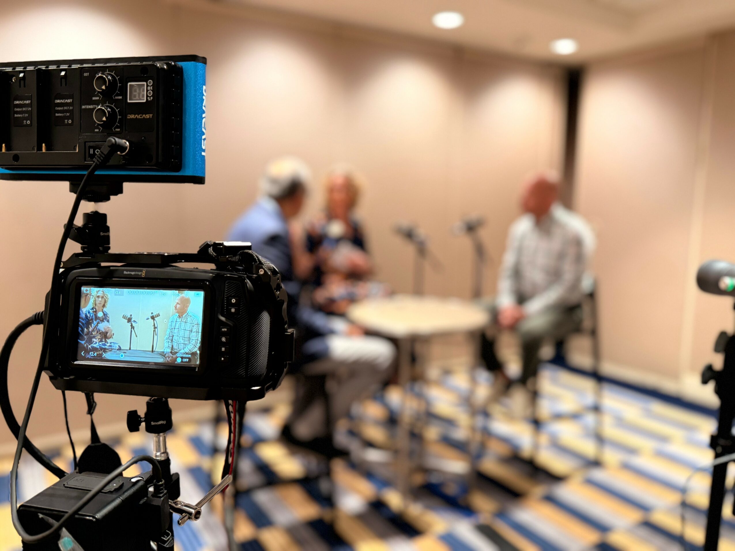 Two people sit at a table with microphones, blurred background, camera in foreground recording the scene, patterned carpet floor.