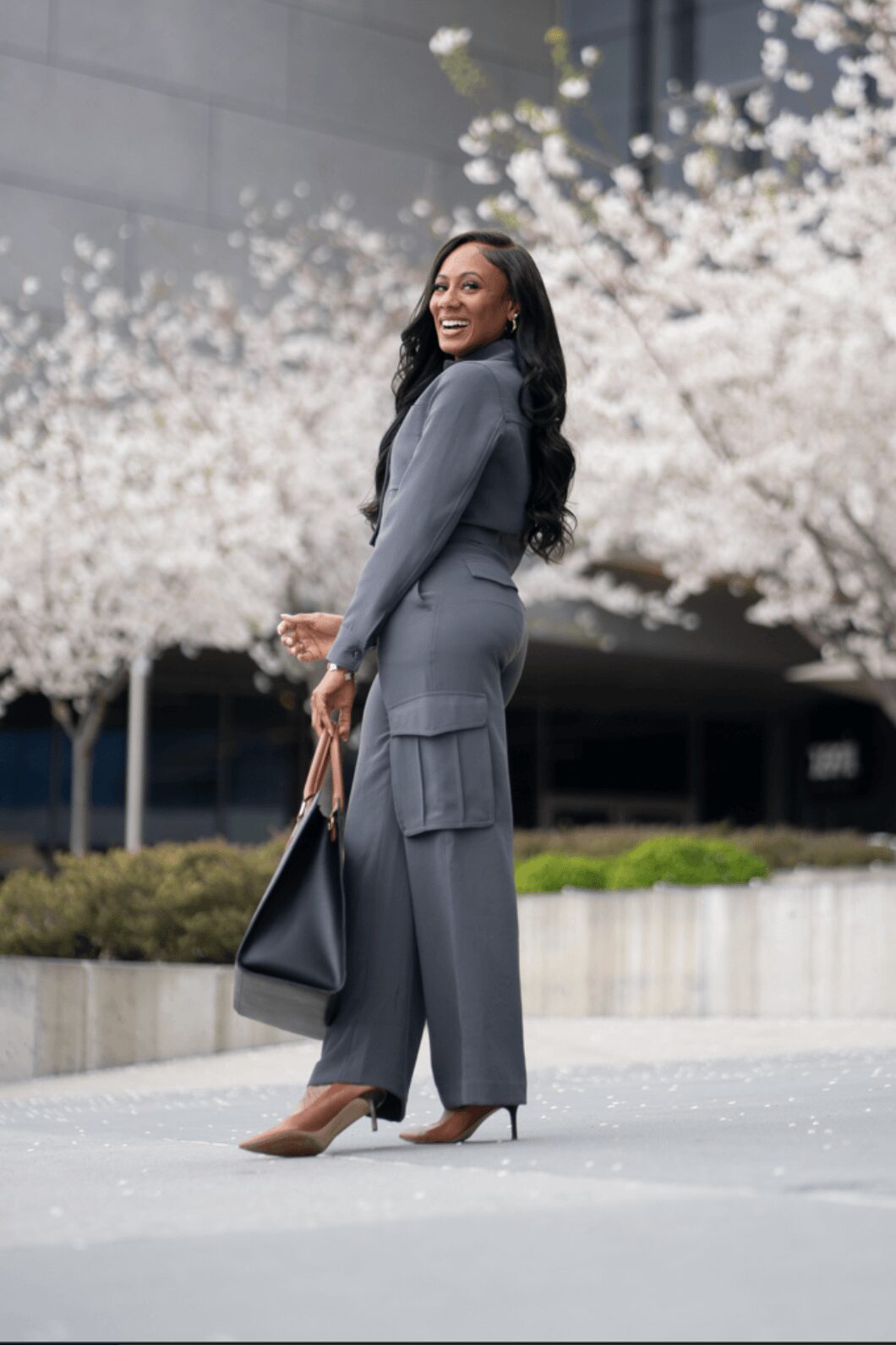 Woman in gray suit holding a black bag, smiling outdoors with blooming trees in background.