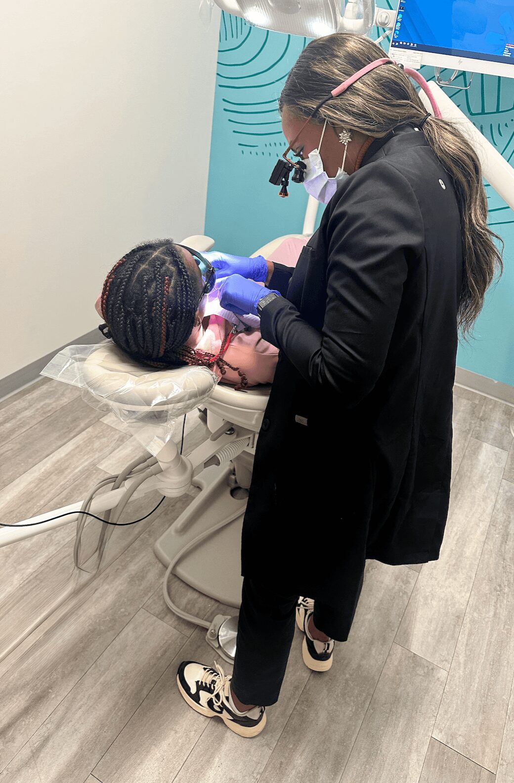 Dental professional examining a child's mouth in a clinic, wearing gloves and a mask, with dental tools and equipment nearby.