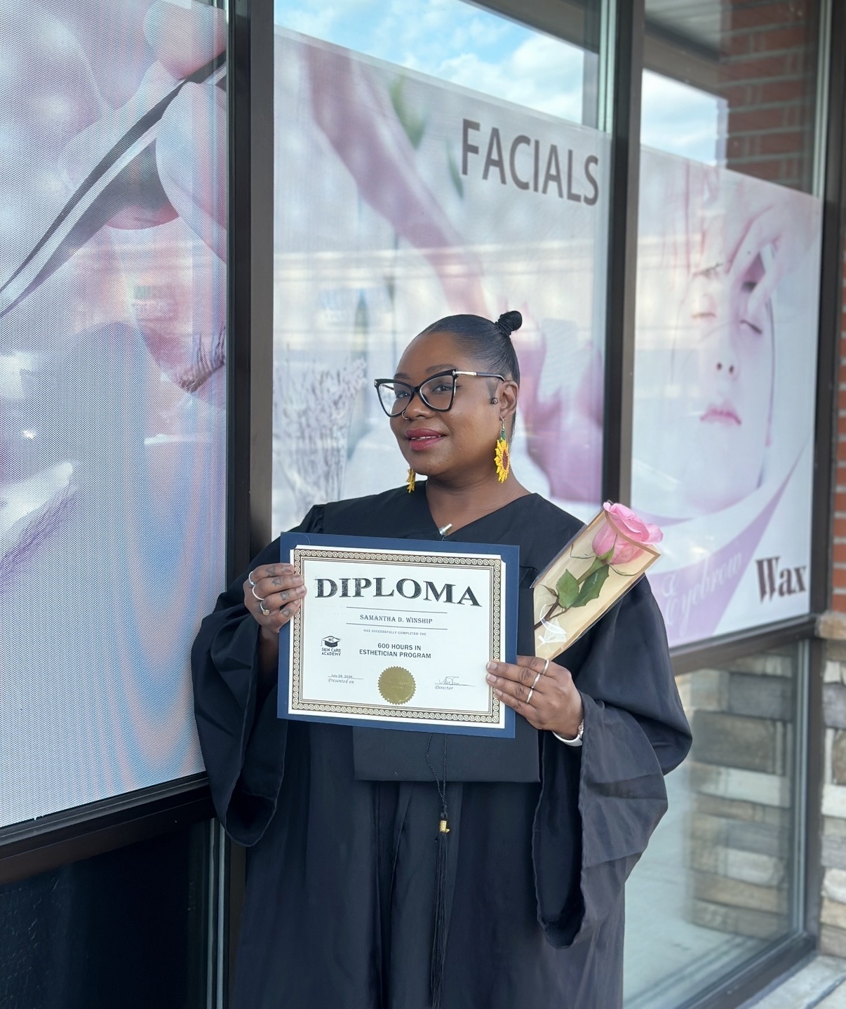 Woman in graduation gown holding diploma and pink rose outside building with facial treatments advertisement, smiling, wearing glasses, earrings, bun hairstyle.