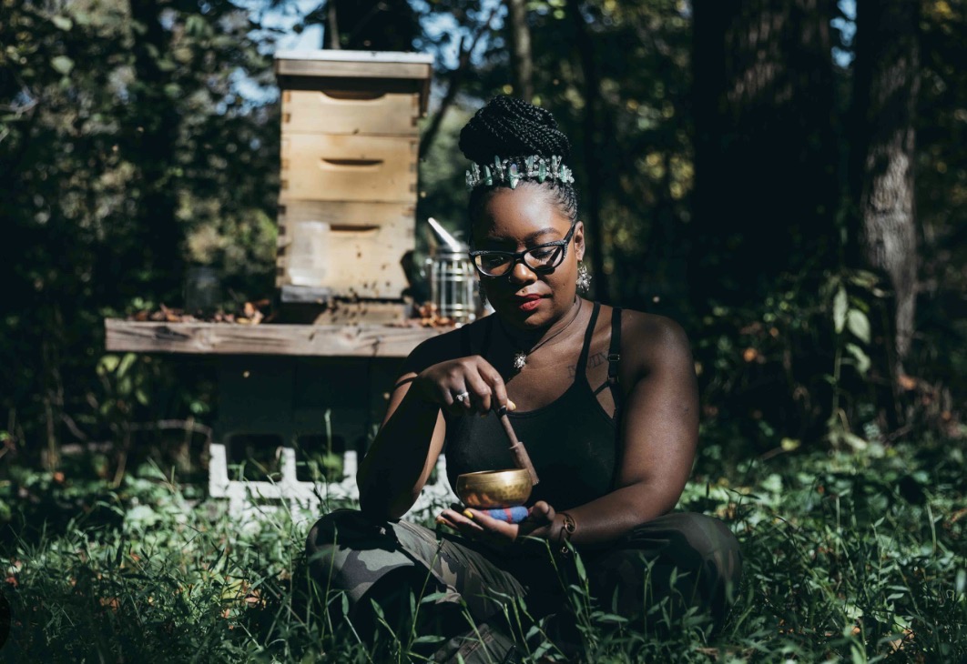 Woman with glasses and headband sitting outdoors, holding a jar, with a beehive and trees in background.