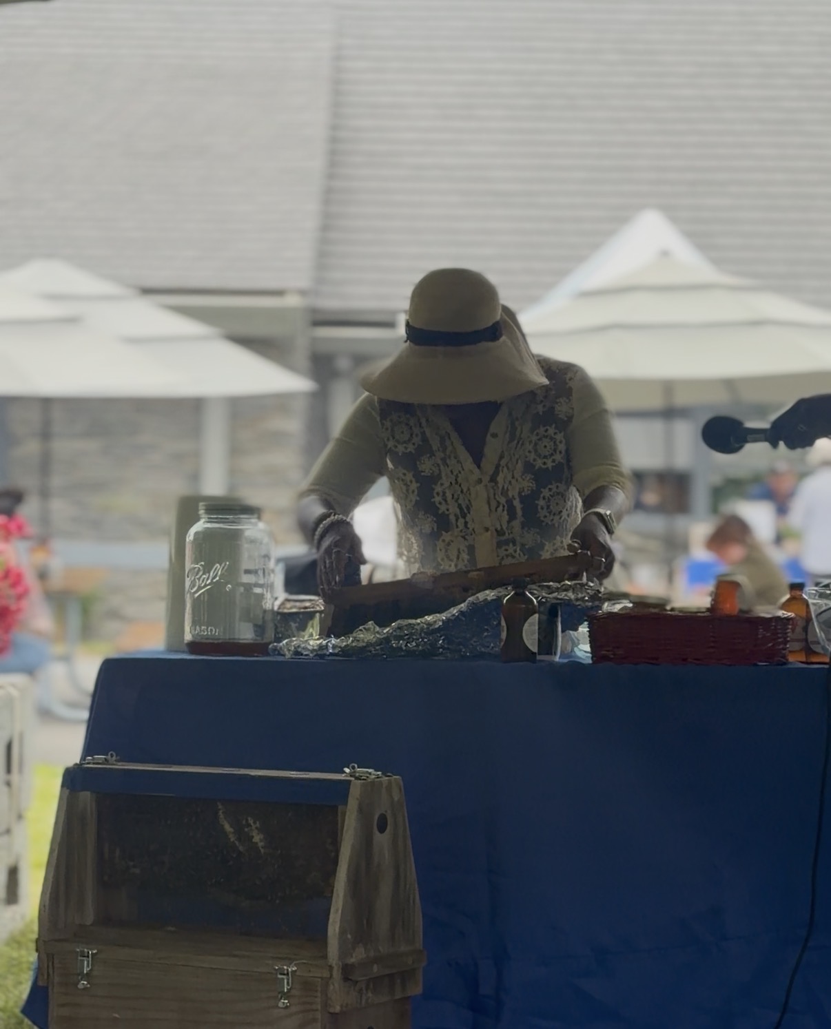 Person wearing a hat and patterned vest preparing food at an outdoor market stall, with umbrellas and people in background.