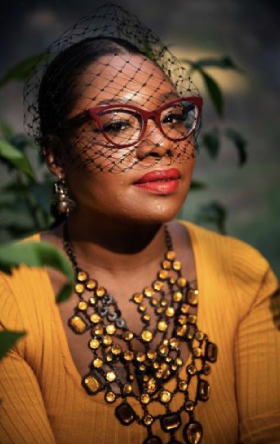 Woman wearing red glasses, yellow top, and layered jewelry, with a black netted veil and greenery background.