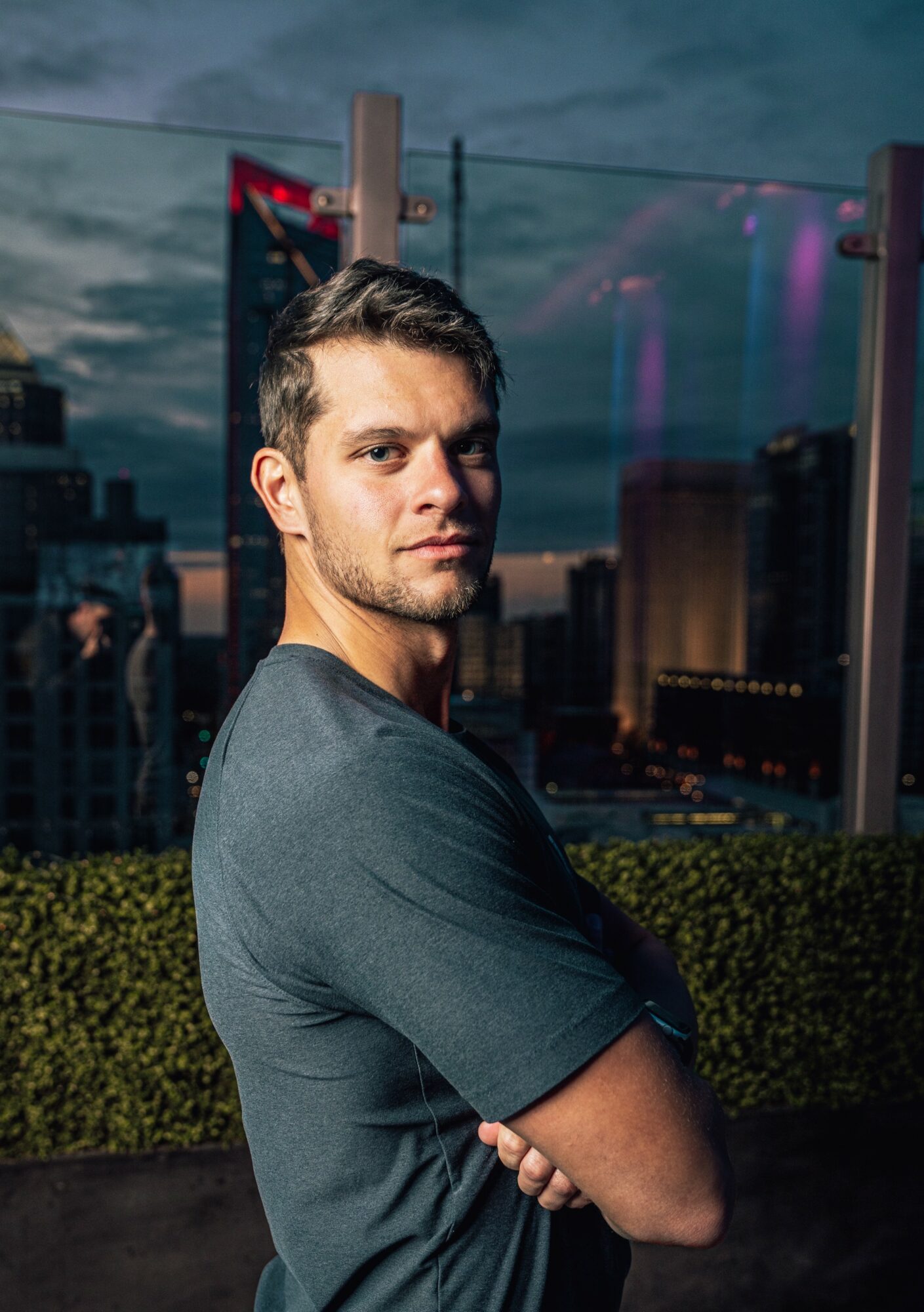 Young man with dark hair and a beard, wearing a gray T-shirt, standing outdoors during evening with city skyline behind him.