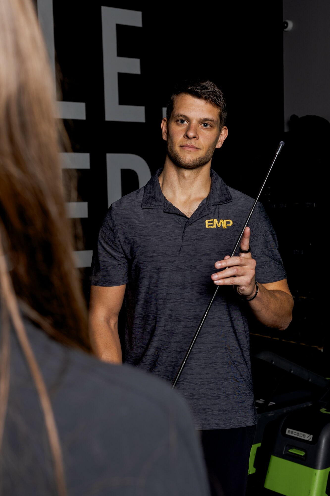 Young man holding a cue stick, wearing a dark polo shirt with 'EMP' logo, standing indoors with a black background.