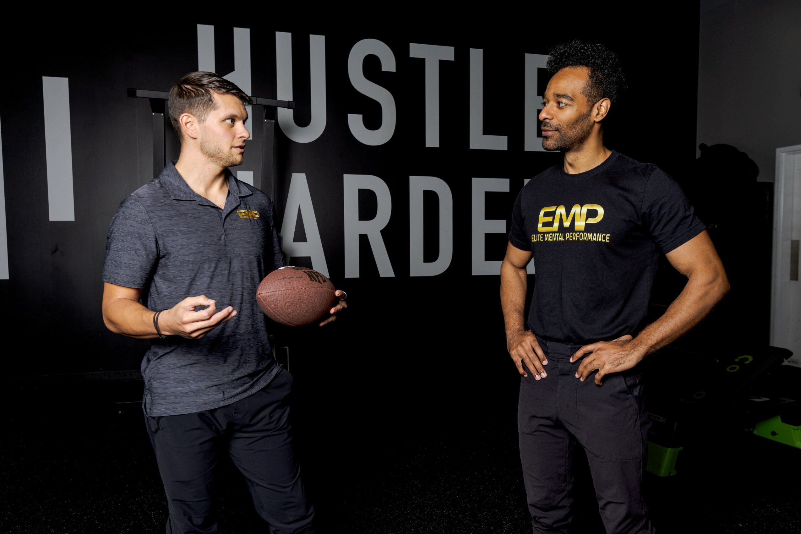 Two men standing and talking in a gym, with a large sign behind them reading 'HUSTLE HARDER'.